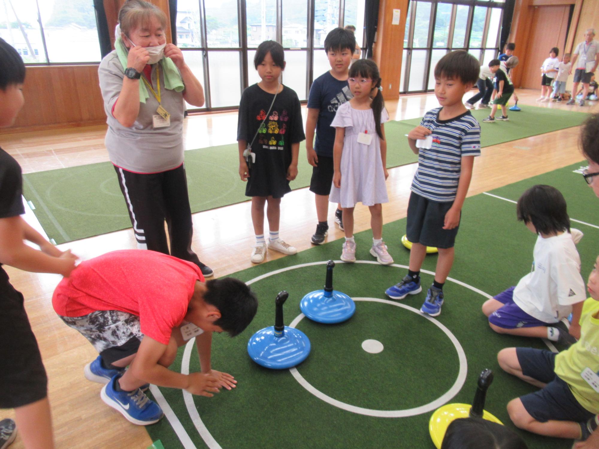 体育館のようなフローリングの室内で、緑色のマットの上でニュースポーツのフロアカーリングのようなアクティビティが行われ、数人の小学生ほどの子どもたちが青いディスクの近くにしゃがみ込み、マスクをした大人がそれを見守っている活動風景の写真