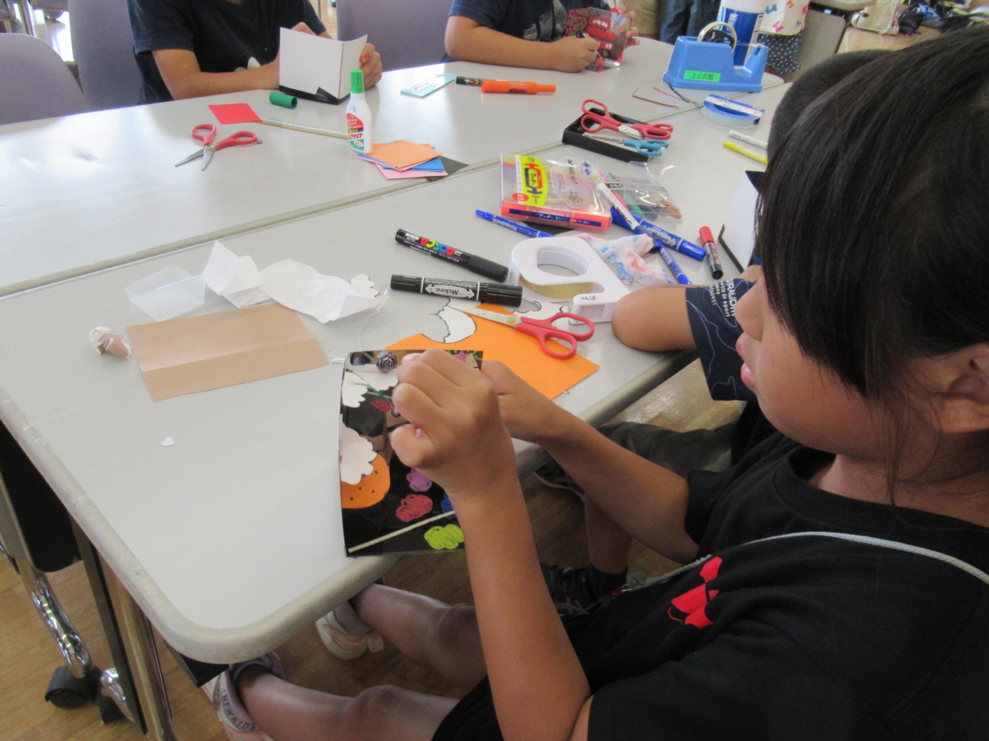 明るい室内の白い長机の上で、色紙やハサミ、のりなどの工作道具が散乱する中、一人の小学校低学年ほどの女の子が集中して黒い台紙にオレンジや黄色の丸い形を貼り付けている、ものづくりワークショップの活動風景の写真