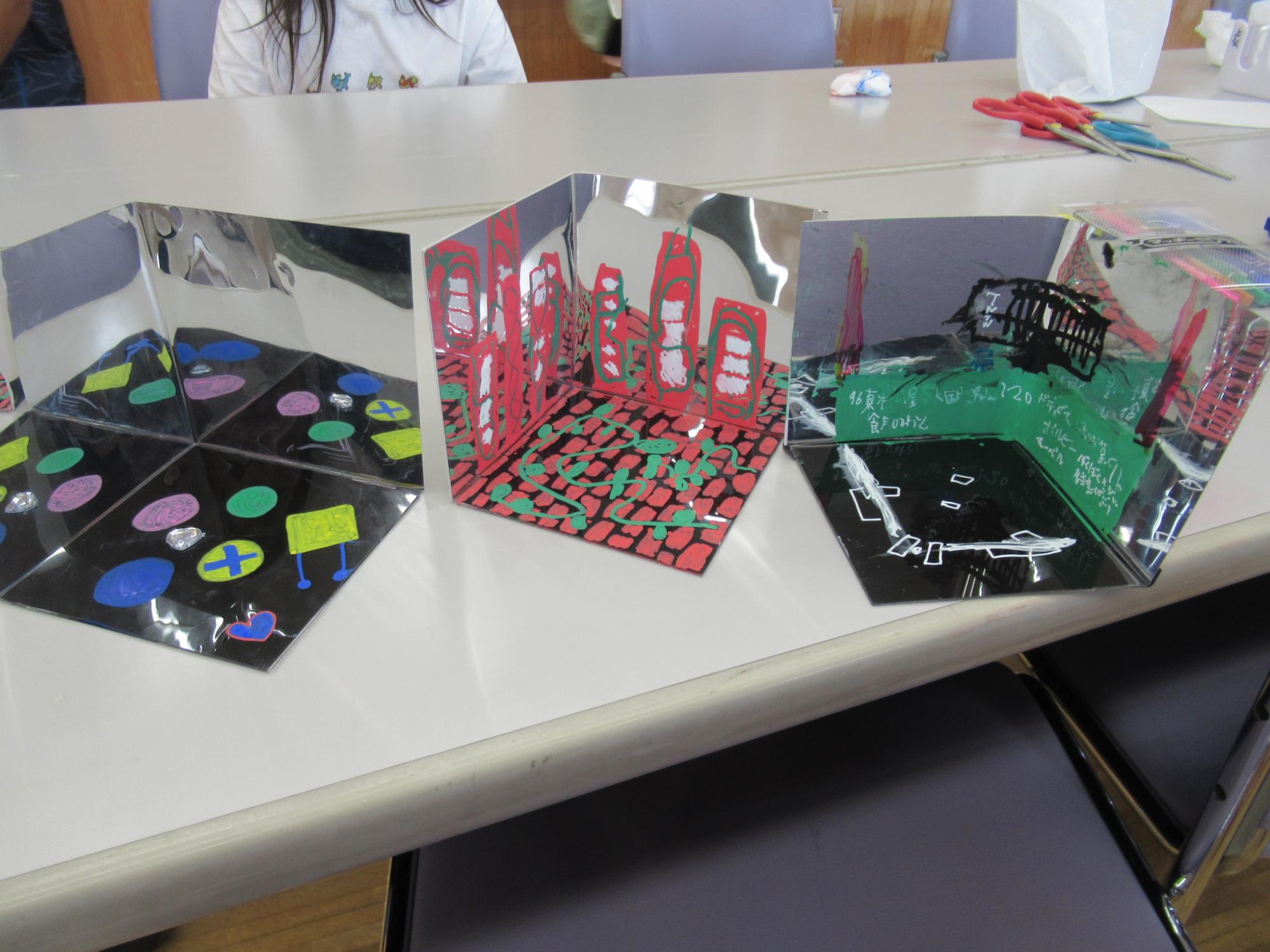 白いテーブルの上に、ミラーペーパーを組み合わせて立体的な空間を作り出し、色付きのシールや切り紙、マーカーなどで都市の風景や抽象的な模様が描かれた3つの万華鏡アート作品が展示されている、ワークショップの様子の写真