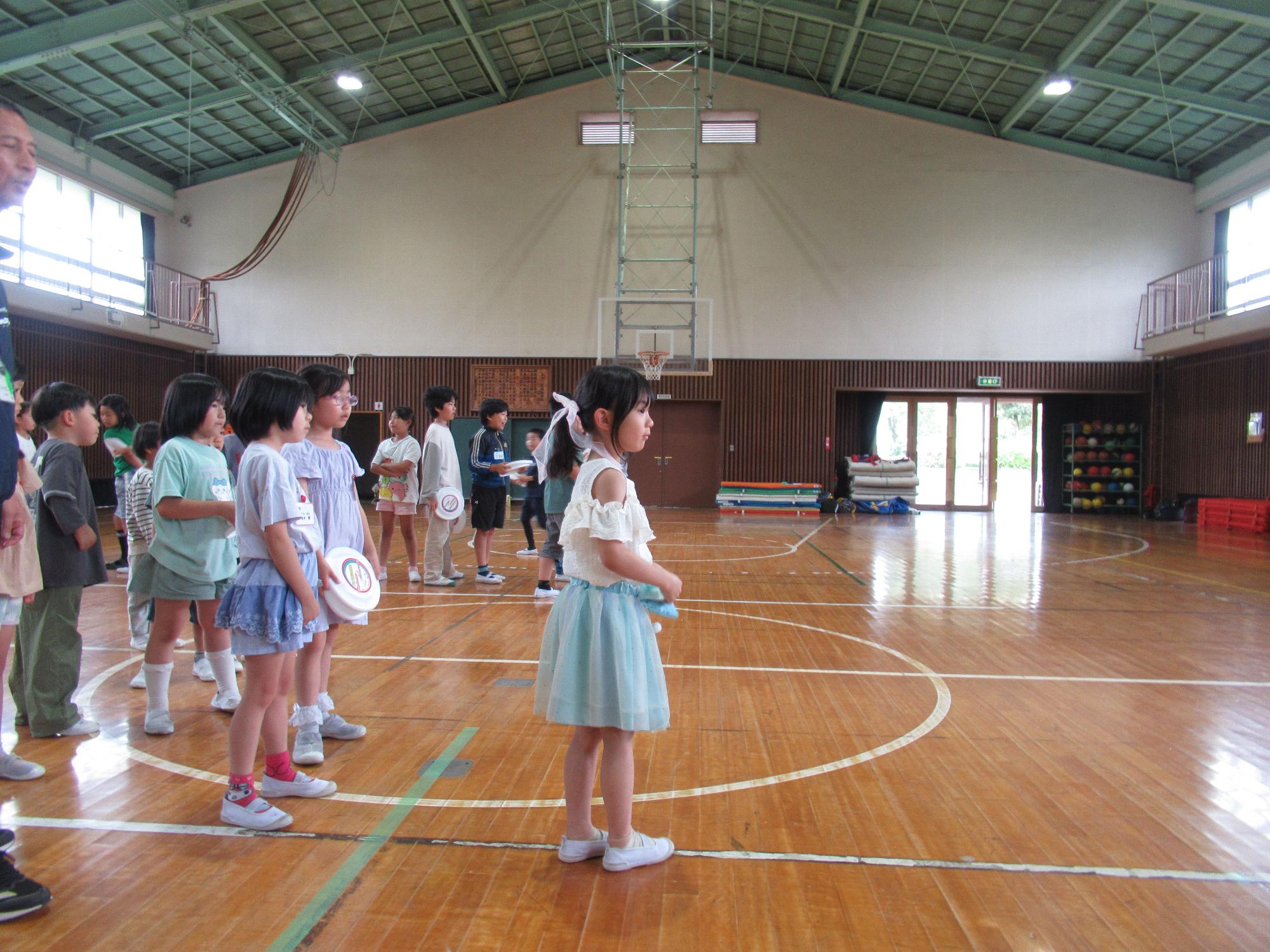 木目調の床の体育館で、小学校の児童と思われる多くの子供たちが横一列に並んでおり、特に水色のスカートを着た女の子が正面を向き、何か活動の指示を待っている様子の写真