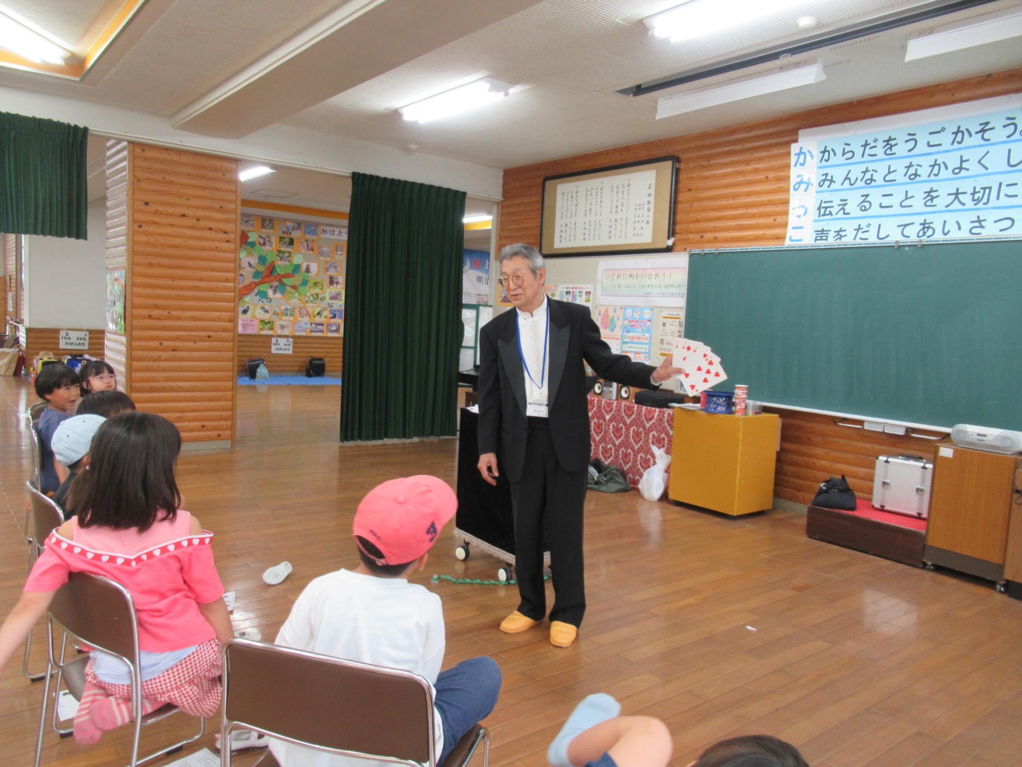 フローリングの広間の中央で、タキシードのような黒いスーツを着た男性がトランプのカードを広げ、椅子に座っている小学校低学年と思われる約10人の子どもたちの前でマジックを披露しているイベントの様子の写真