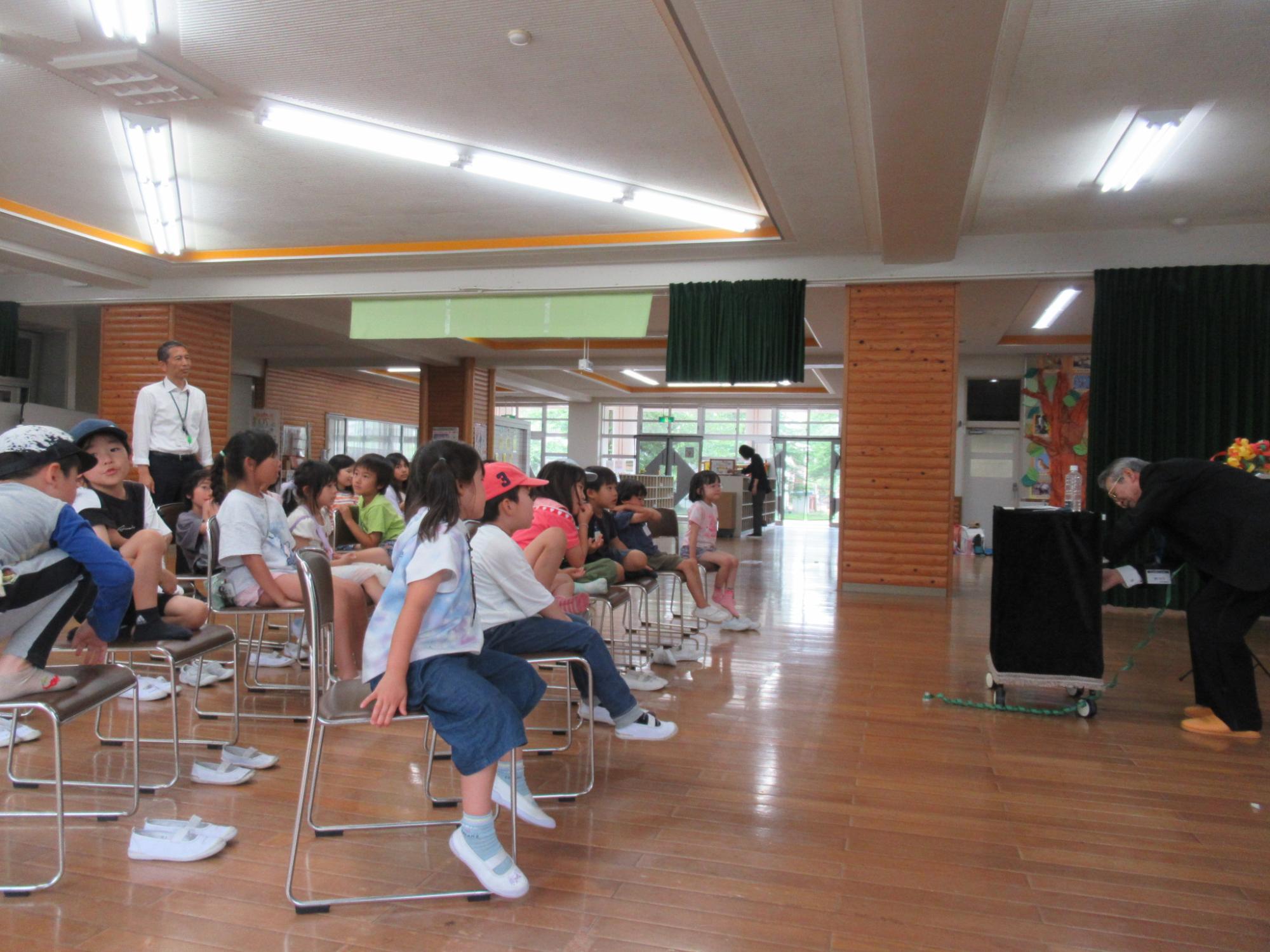 フローリングの広い室内で、黒いスーツ姿の男性が黒い布のかかった台を使ってマジックを披露しており、それを見物する約20人の小学生ほどの子どもたちが、パイプ椅子に座って真剣な表情や楽しそうな表情で見入っているイベントの様子の写真