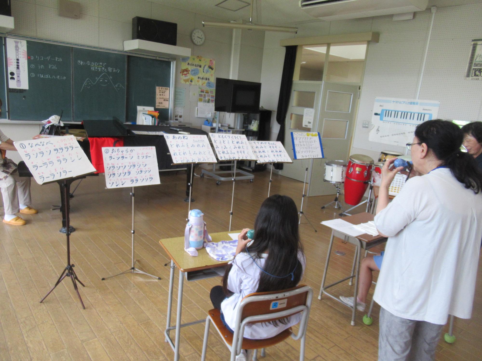 教室のような室内で、黒板の前にティンパニやドラムセットなどの楽器が置かれ、譜面台には歌詞と音階が書かれた紙がクリップで留められており、一人の女の子が机に向かい、女性が写真を撮っている音楽活動の準備風景の写真