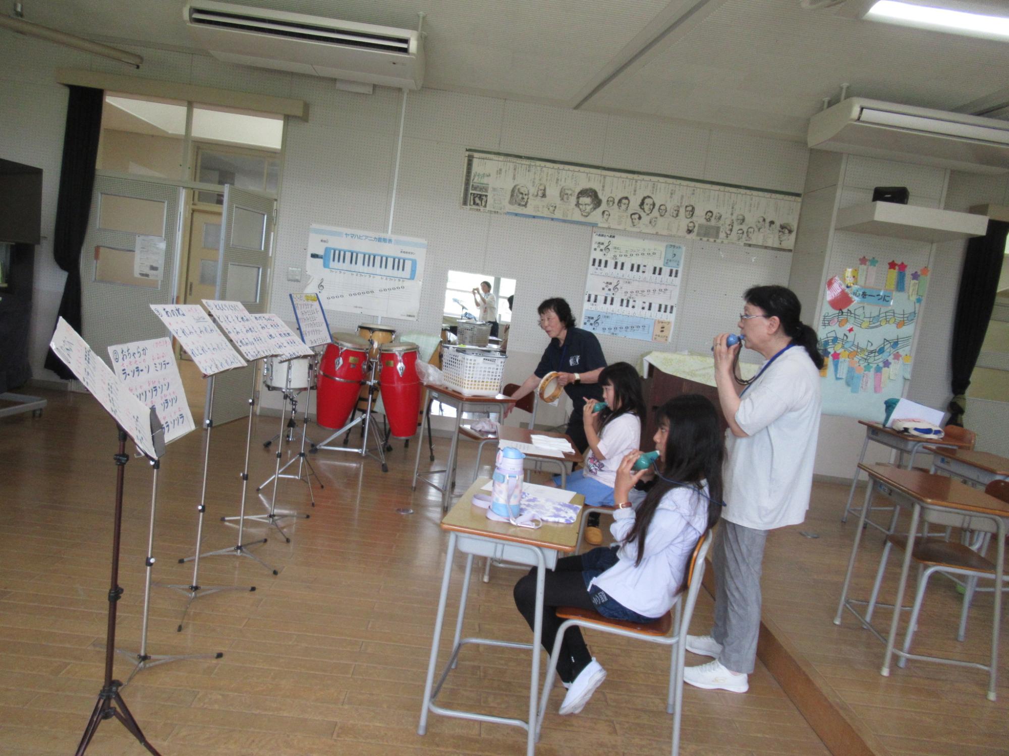 音楽室のような室内で、椅子に座った2人の女の子の間に女性が立ち、それぞれオカリナを構えており、譜面台に立てかけられた楽譜を見ながら練習風景の写真