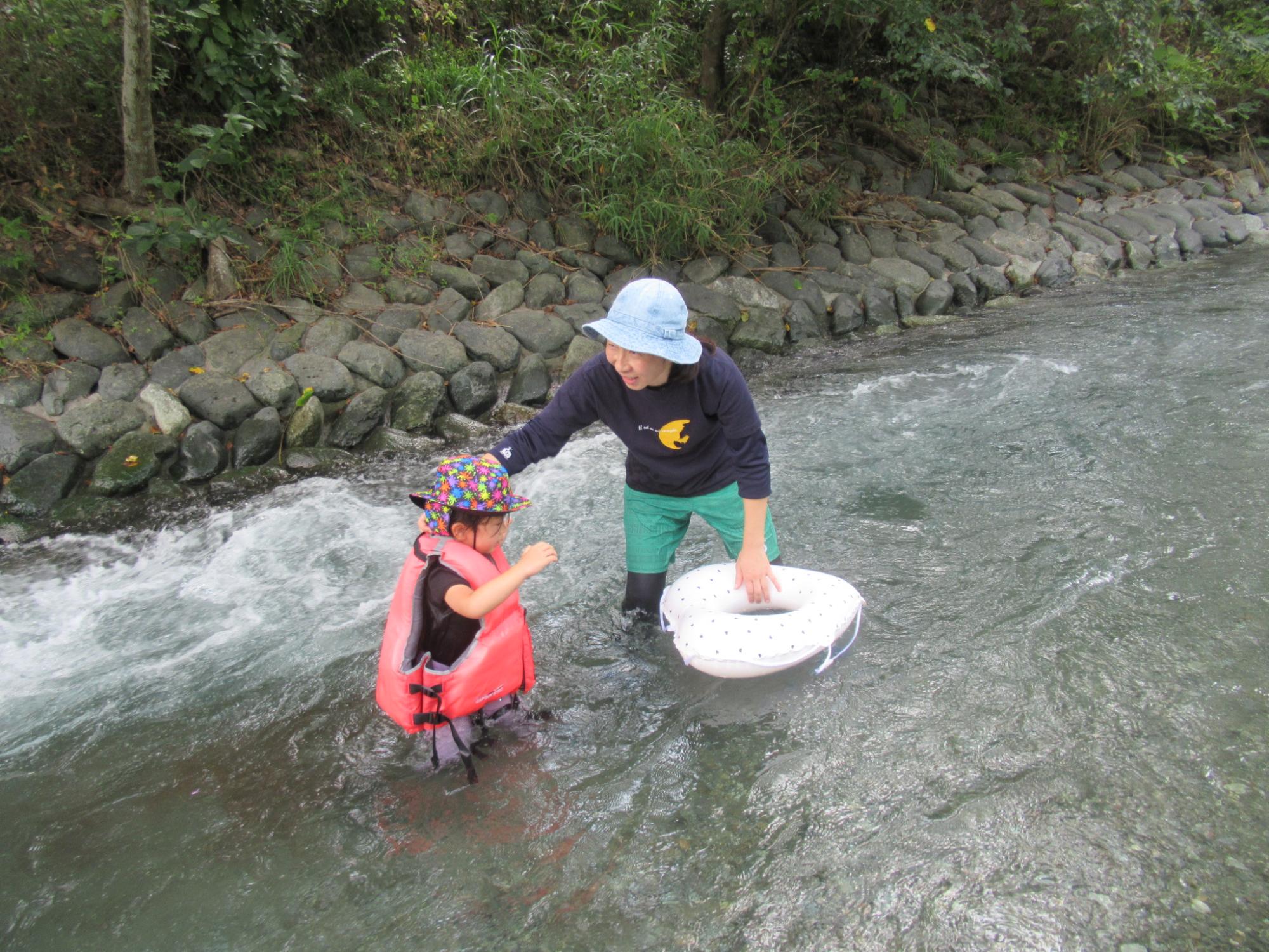 ライフジャケットを着て水に浸かる子どもに、水色の帽子をかぶった大人が寄り添い、白い水玉模様の浮き輪を持って浅い川で水遊びを教えている様子の写真