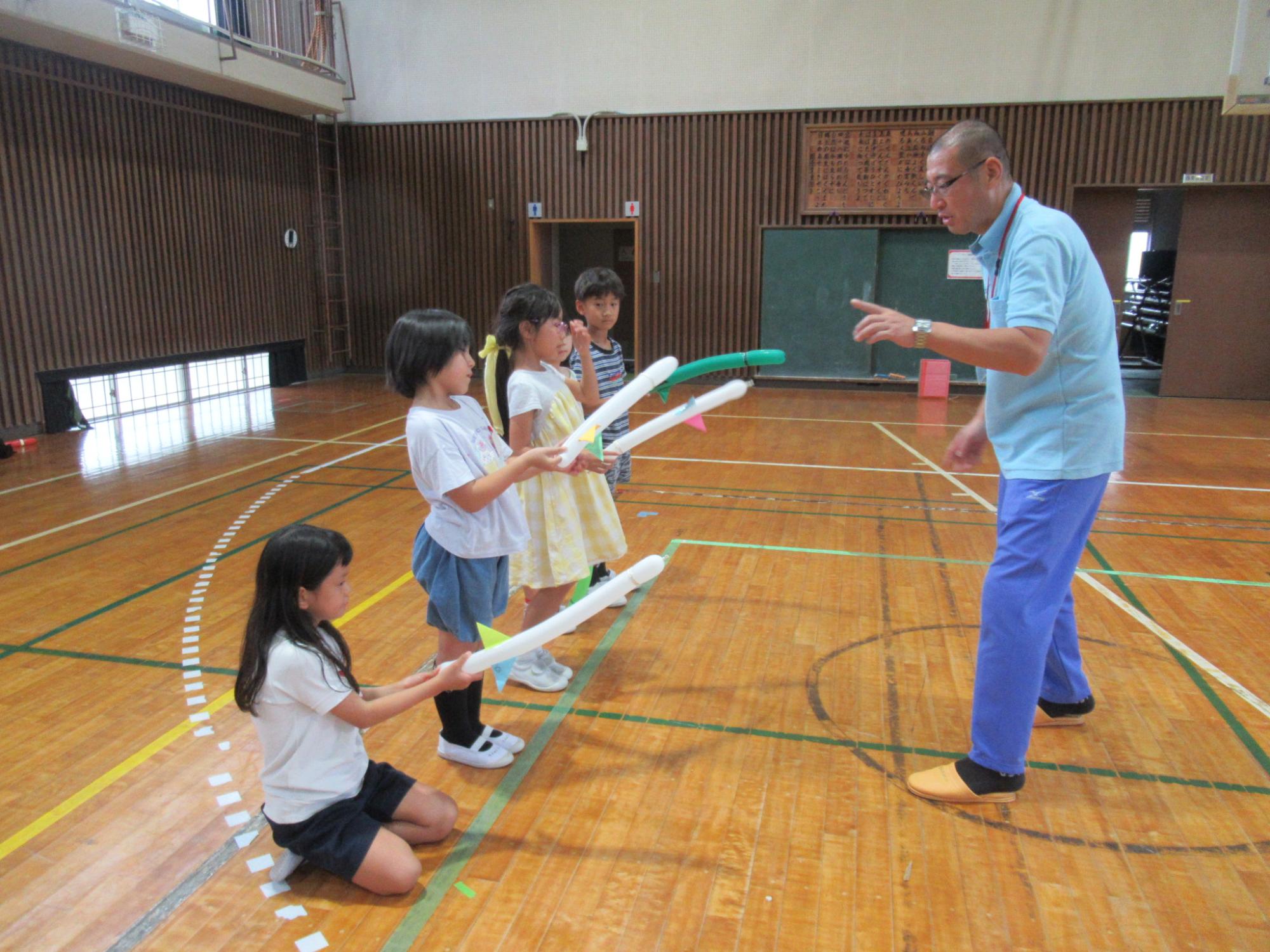 体育館のフローリングの上で、水色のポロシャツと青いズボンを履いた男性が、バルーンソードの細長い風船を持った数人の小学校低学年ほどの子供たちに、バルーンアートの説明している写真