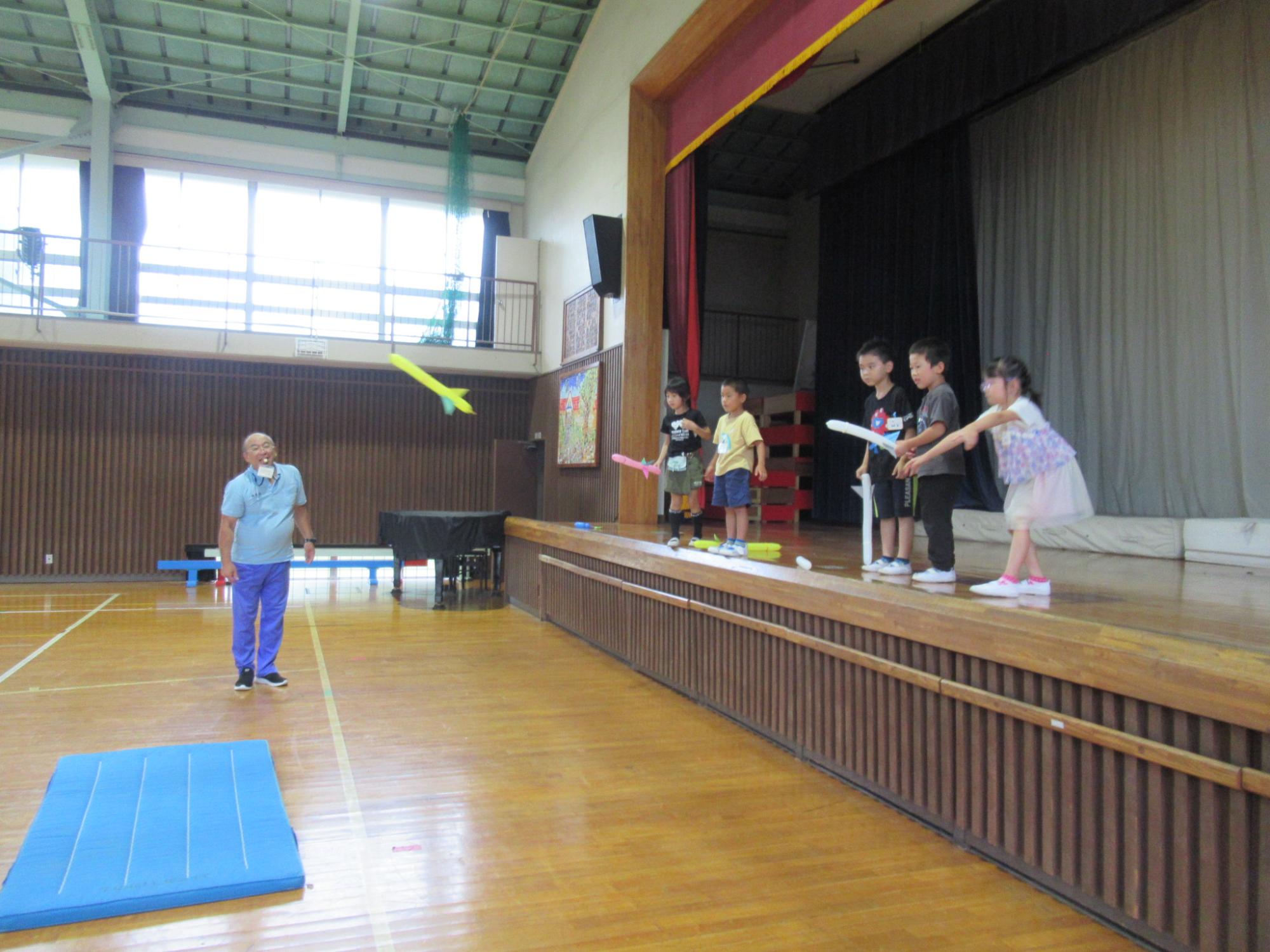 体育館のステージ上から、細長いバルーンソードの風船を持った数人の小学生ほどの子どもたちが、ステージ下のマットに向かって、風船を投げている写真