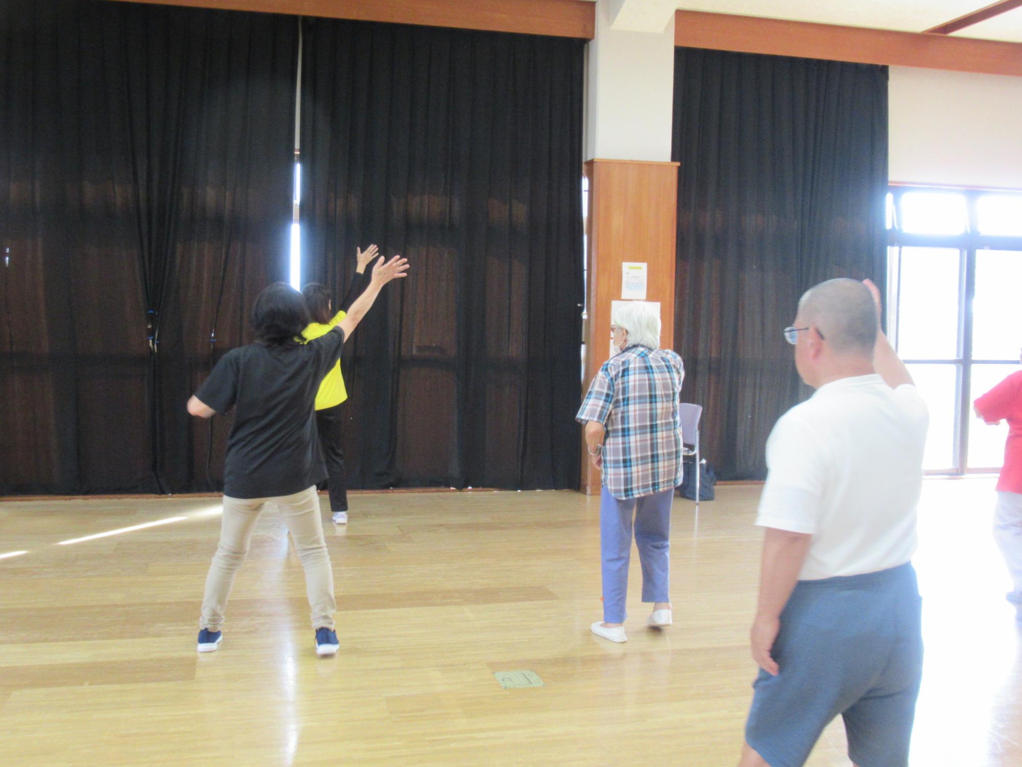 黒いカーテンの引かれた体育館で、数人の高齢者が腕を大きく振り上げたり、体を動かしたりして健康体操やレクリエーションを楽しんでいる様子の写真