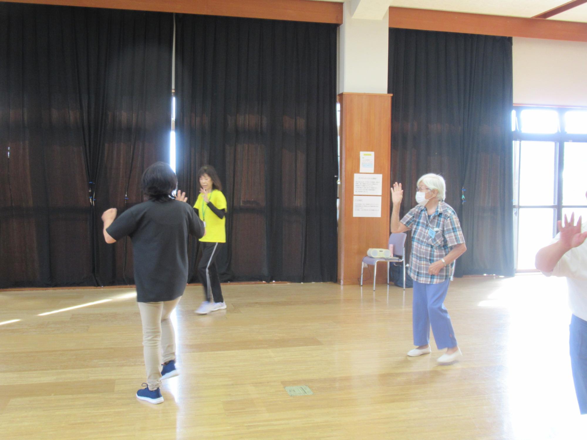 数名の高齢者が黒いカーテンが引かれた体育館で、インストラクターらしき人物に合わせて手を上げたり体を動かしたりする軽運動や体操に取り組んでいる様子の写真
