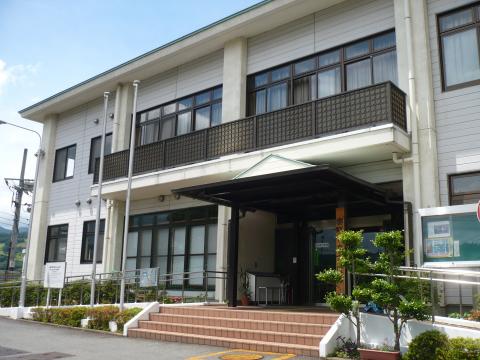 二階建ての白い建物正面に、屋根付きの入口と植栽、スロープなどが整備されている上公民館外観写真