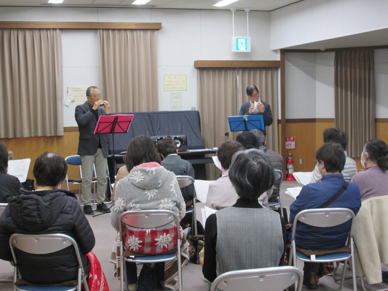 オカリナとバンジョーを演奏する男性2人と、資料を手に演奏を楽しんでいる参加者たちの写真