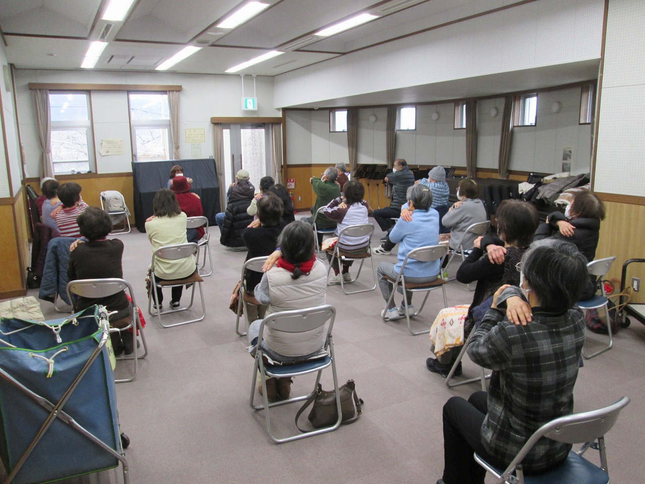 輪になって椅子に座った高齢者が、講師の指示に従い、両腕を交差させて肩を揉むような体操をしている様子を捉えた写真