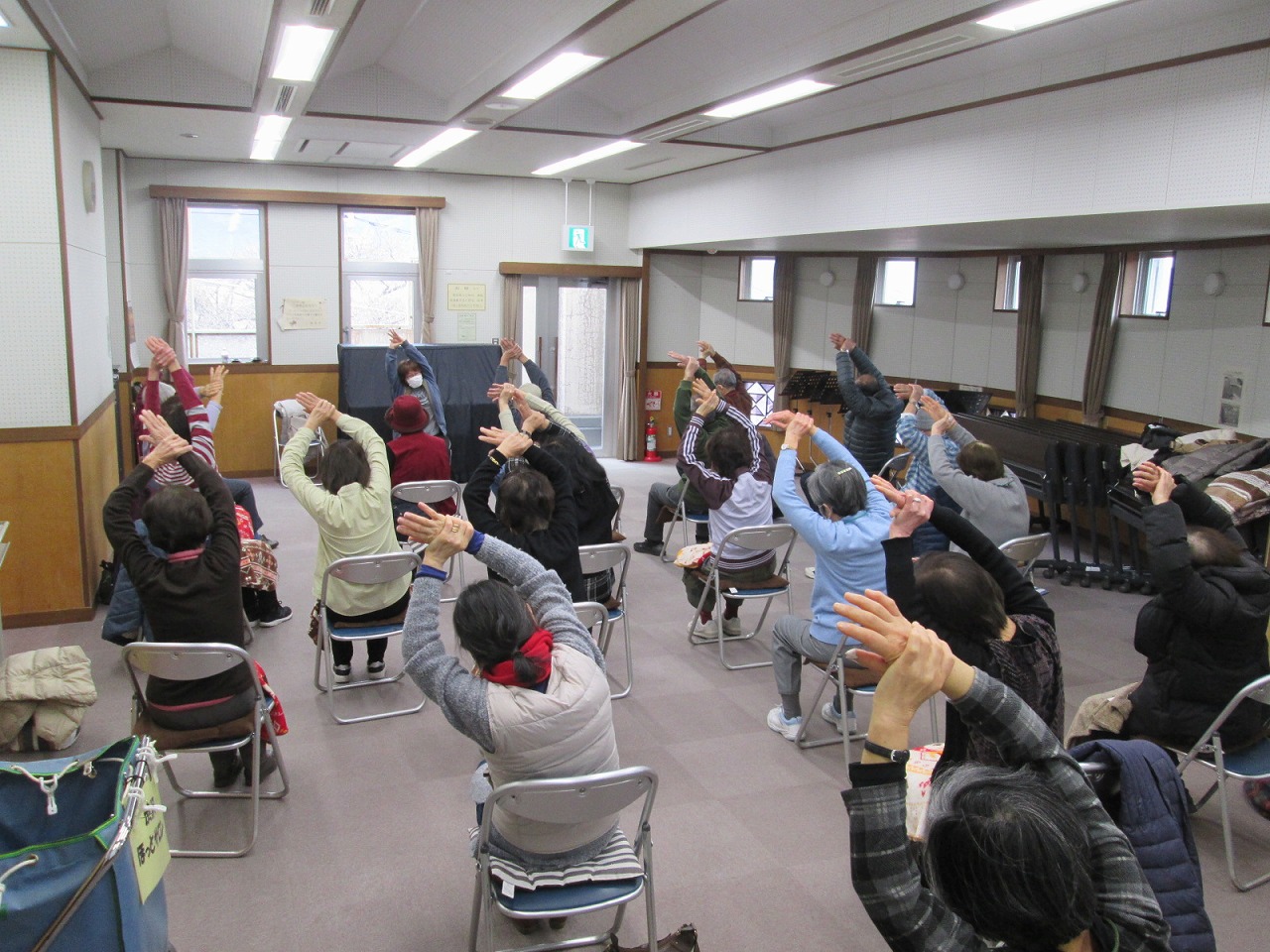 輪になって椅子に座った高齢者が、両腕を上げて横に体を曲げるストレッチをしている様子を捉えた写真