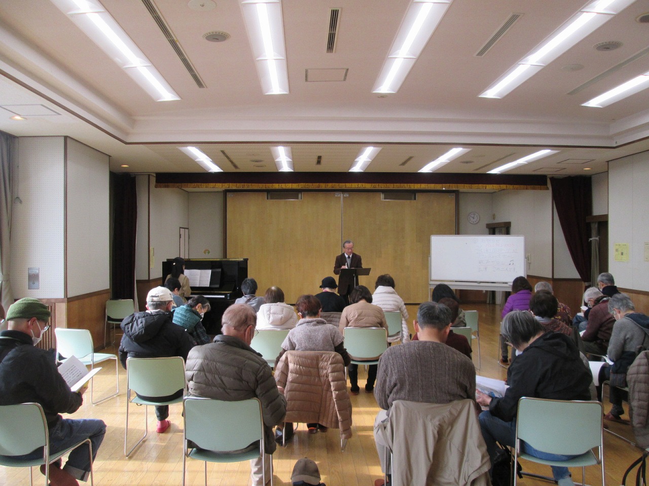 屋内広間に集まった参加者たちが前方の演壇に立つ男性の講演を聴いており、熱心にメモを取っている様子の写真