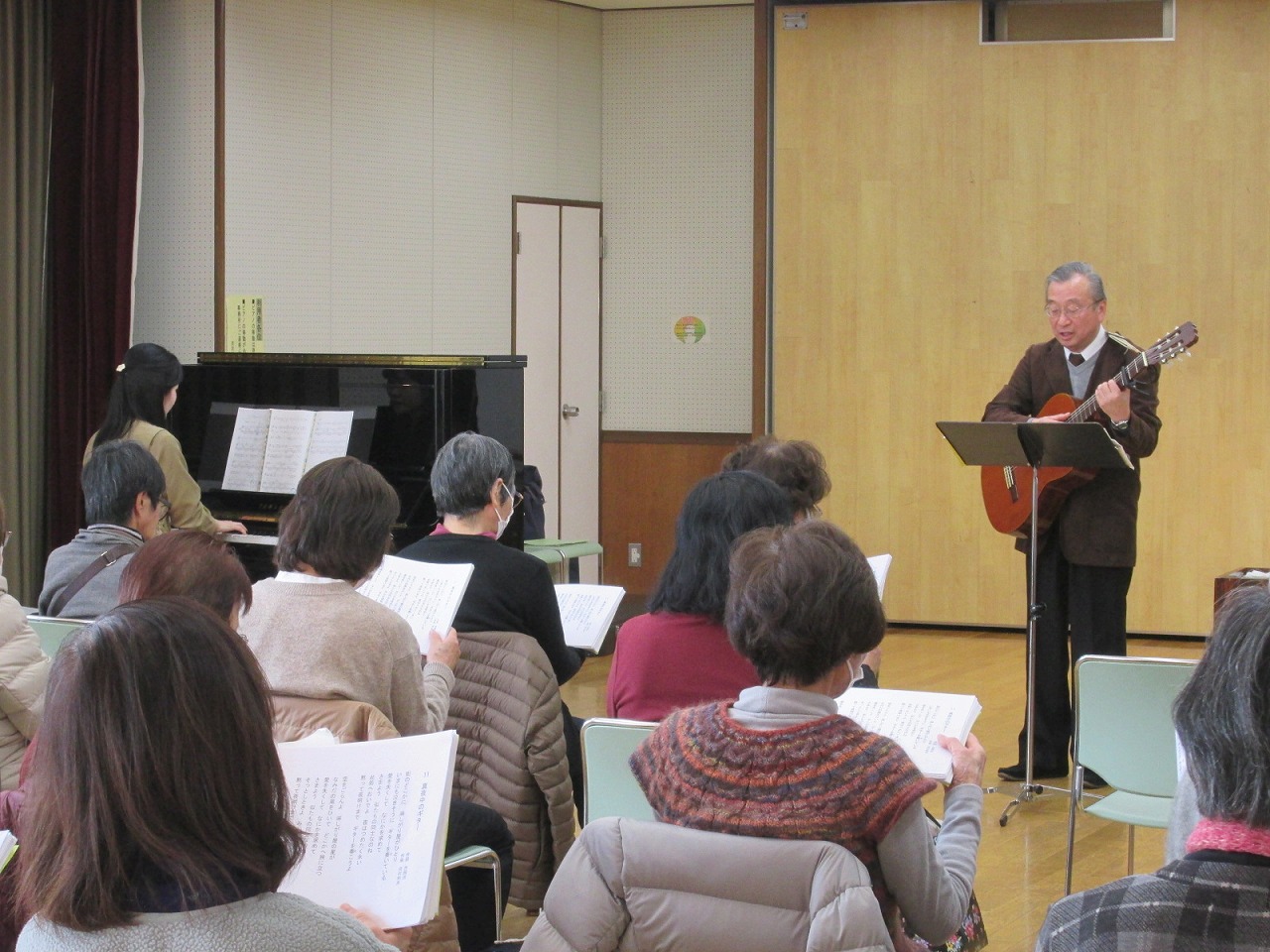 ギターとピアノの伴奏に合わせて、数人が椅子に座って歌詞を見ながら歌っている様子の写真