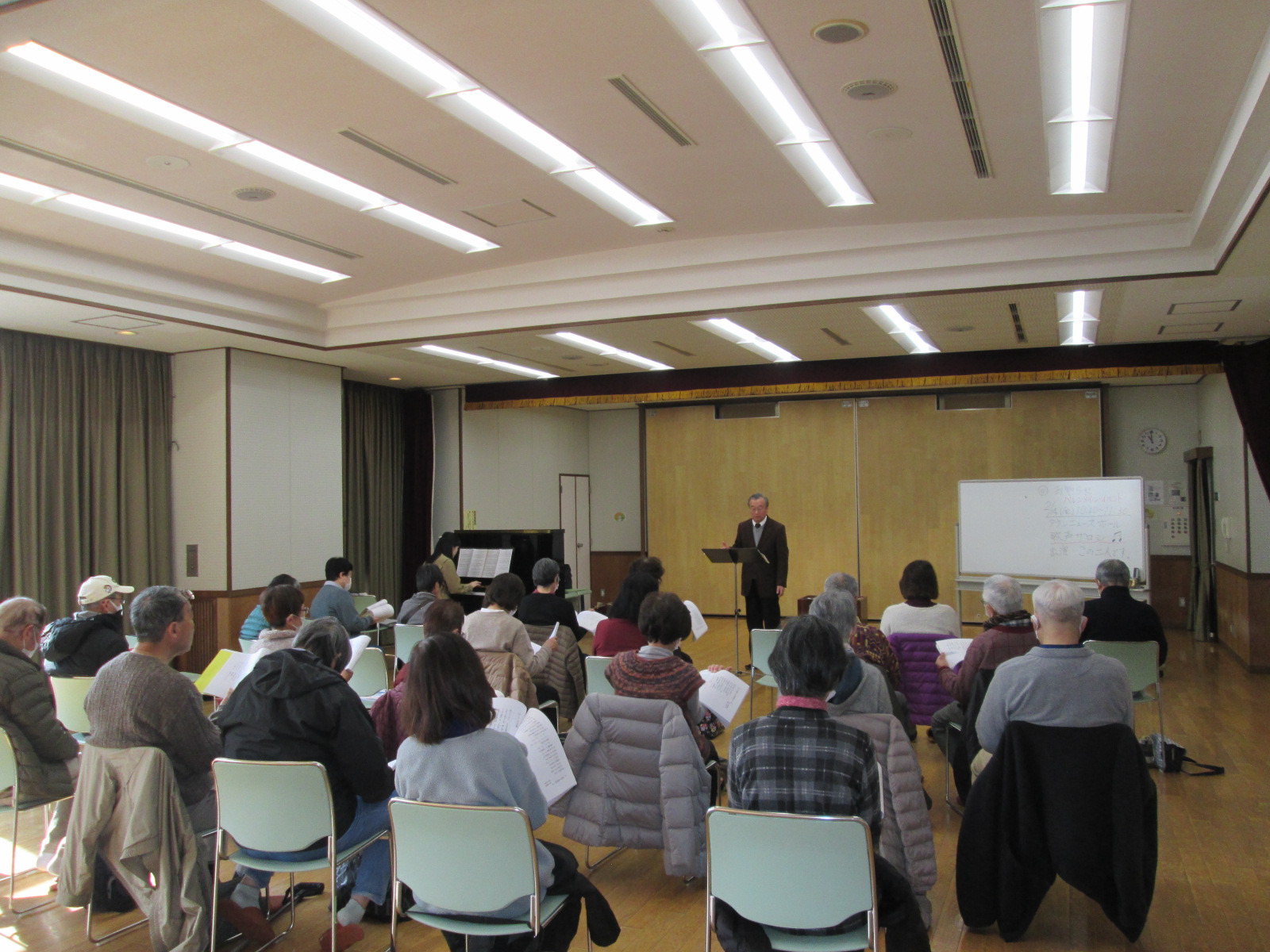 広い部屋で、多くの人が椅子に座ってギターを持った男性の前で合唱練習をしている様子の写真