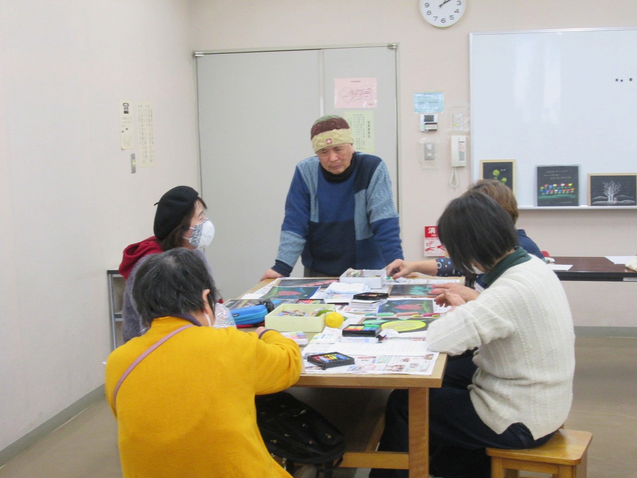 室内の木製テーブルを囲み、帽子をかぶった男性が数名の参加者に対し、テーブルの上に広げられたカラフルな絵本のようなものや道具を使って、何かを熱心に教えている様子の写真