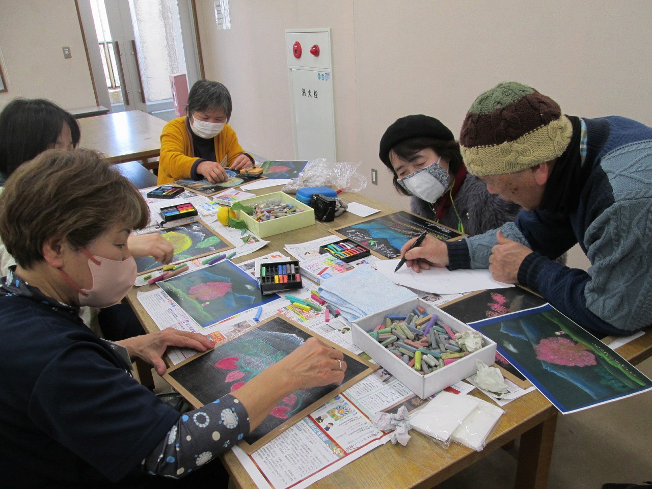 帽子をかぶった年配の男性とマスクを着用した数名の女性たちが、色とりどりのパステルやクレヨンを使い、黒い台紙の上に描かれた花や魚などの絵画作品制作に熱心に取り組んでいる美術教室の様子の写真