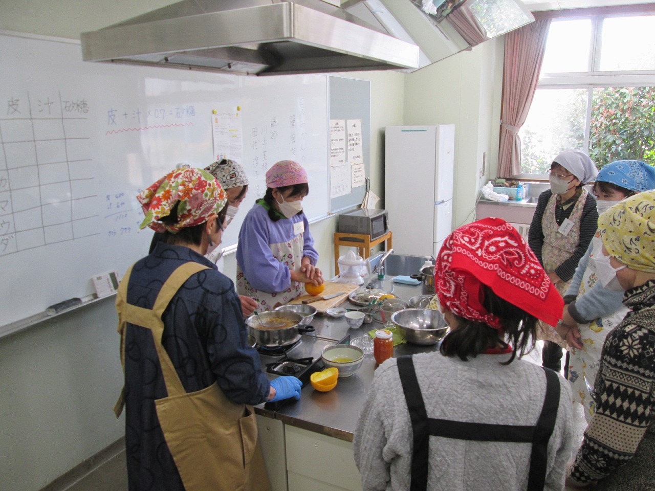 多くのエプロンとバンダナまたは三角巾を着用し、マスクを着けた女性たちが、ホワイトボードのレシピを見ながら調理台の上で柑橘系の果物の皮むきなど調理を行っている料理教室の様子の写真