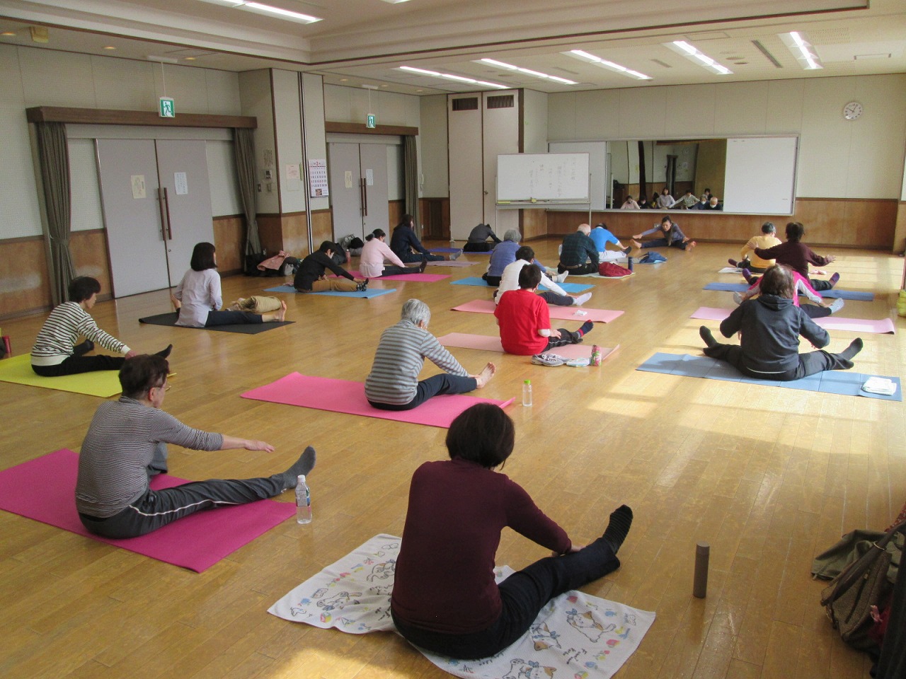 フローリングの広い室内で、様々な色のヨガマットを敷いた十数人の人々が、両足を大きく開いて腰を落とし、前方に身を乗り出すようなポーズで運動に励んでいる様子の写真
