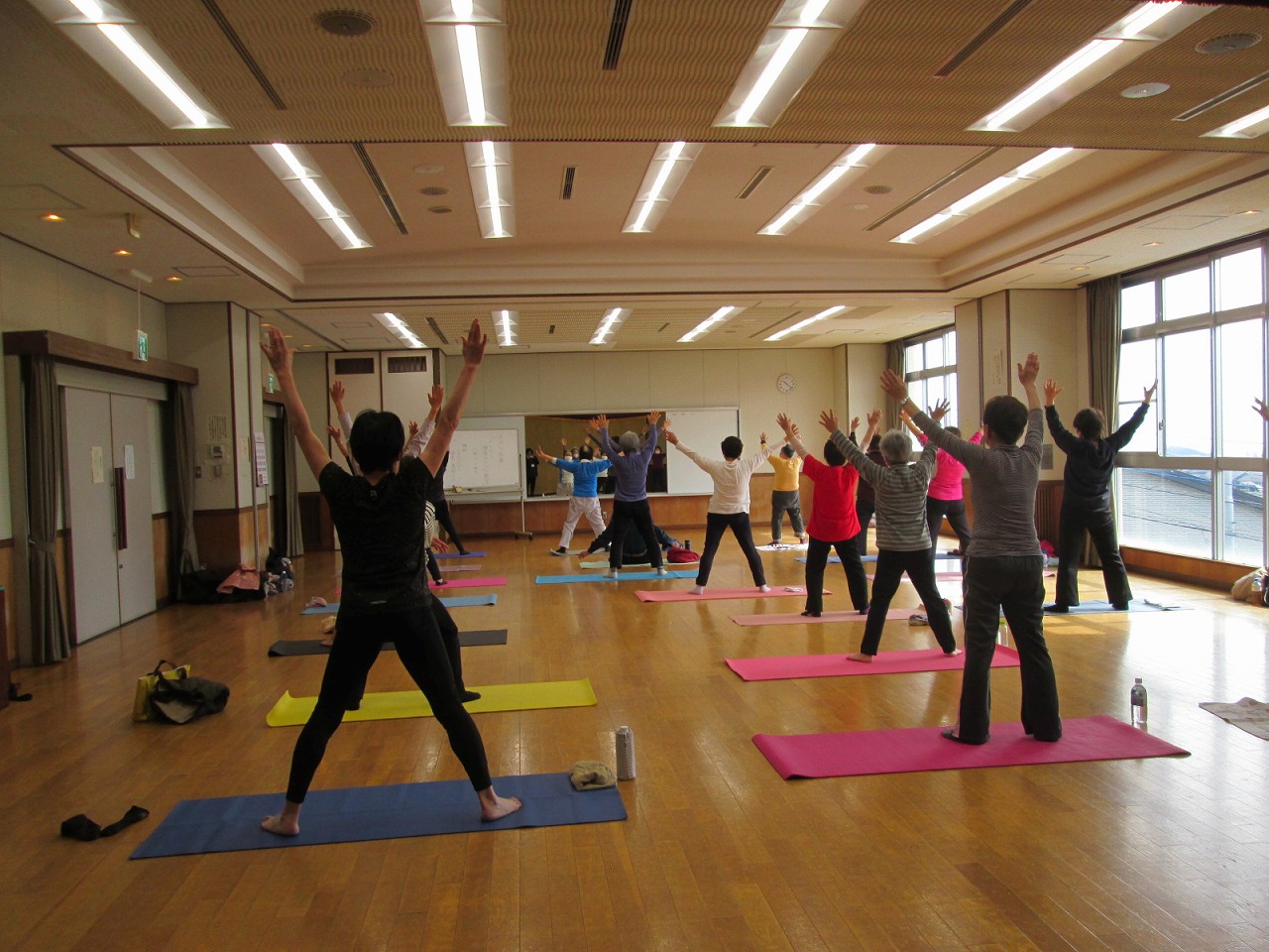 窓からの自然光と天井の照明が明るく照らす中、ヨガマットを敷いた十数人の人々が両手を頭上にまっすぐ伸ばすポーズで体操やヨガに励んでいる様子の写真