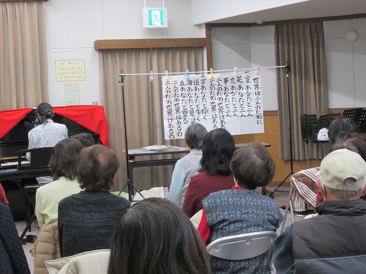 クリップで吊るされた歌詞を前方に掲げ、椅子に座った高齢者の参加者たちが、ピアノを弾く指導者のもとで合唱練習をしている様子を捉えた写真