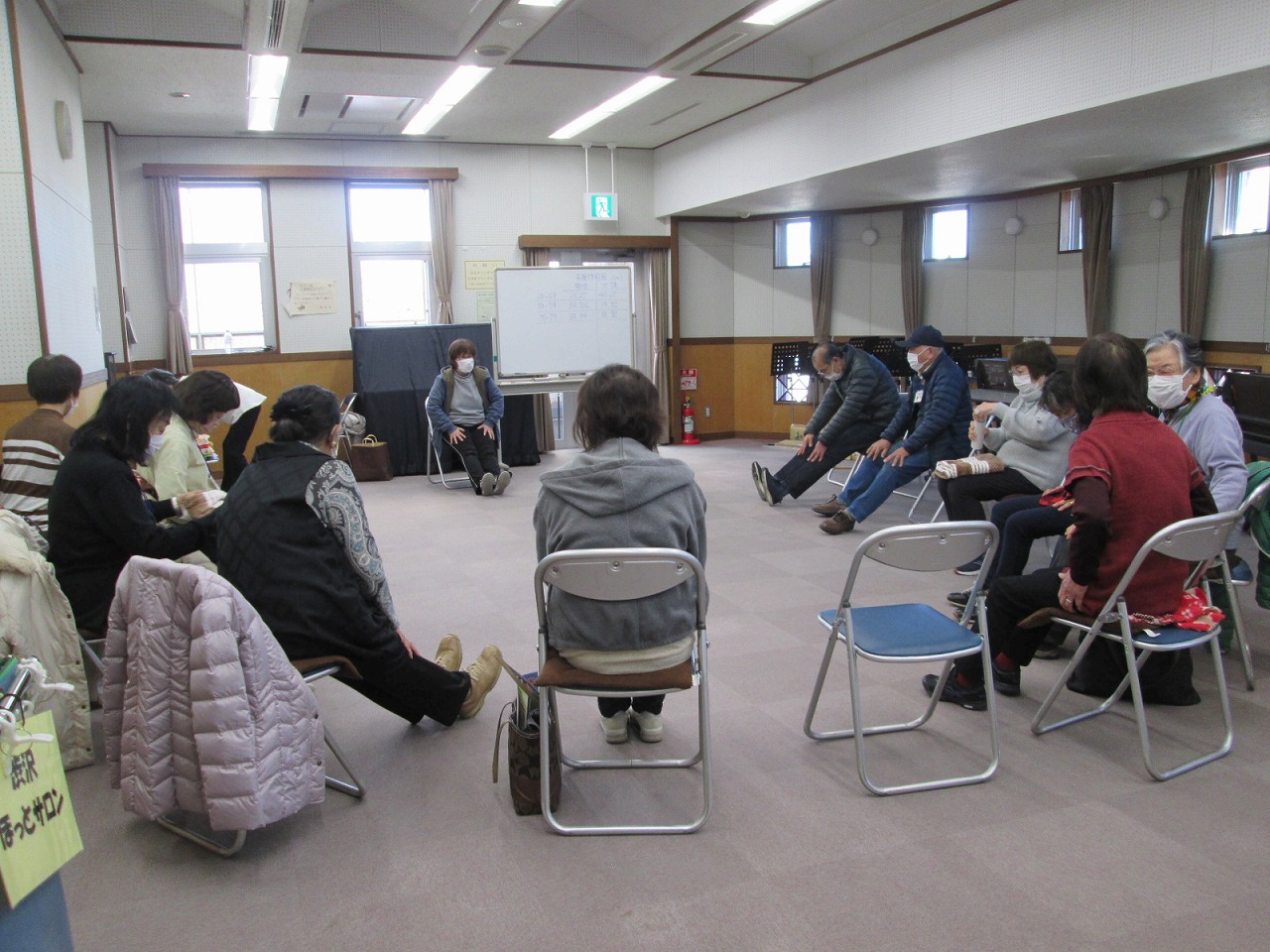 輪になって椅子に座っている参加者たちが、指導者の指示に従って床に足を伸ばす体操をしている様子を捉えた写真