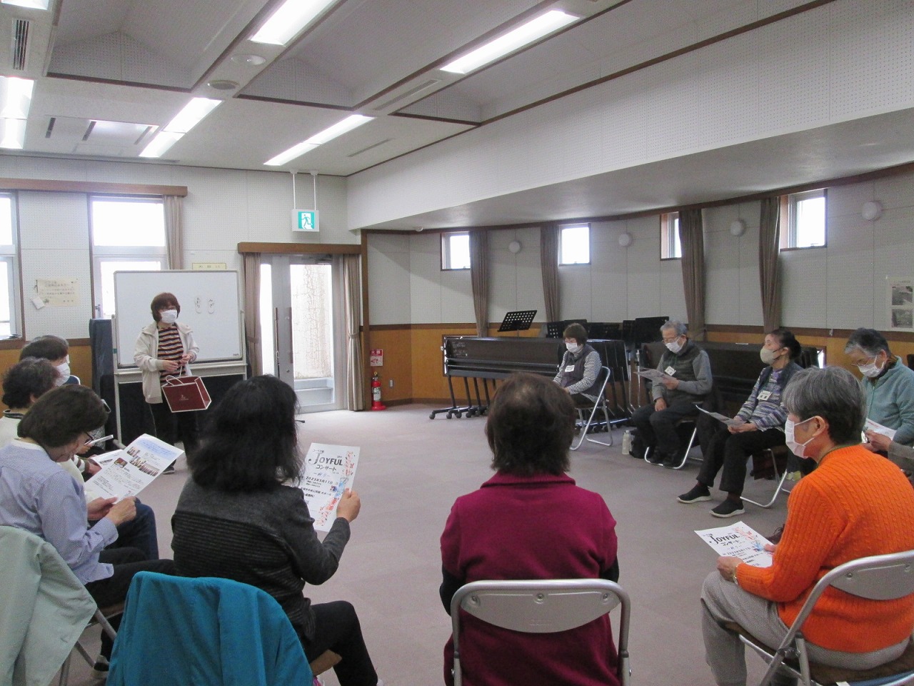 女性が立つホワイトボードを囲んで円を描いて並んだ椅子に座り、資料を広げた参加者たちの写真