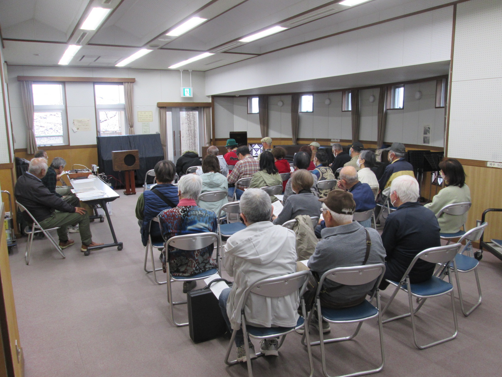 大勢の参加者が集まり、広い室内で前方のテーブルで資料を広げる講師と思われる人物に向かって、青と銀色の椅子に座りながら熱心に話を聞いている様子の写真