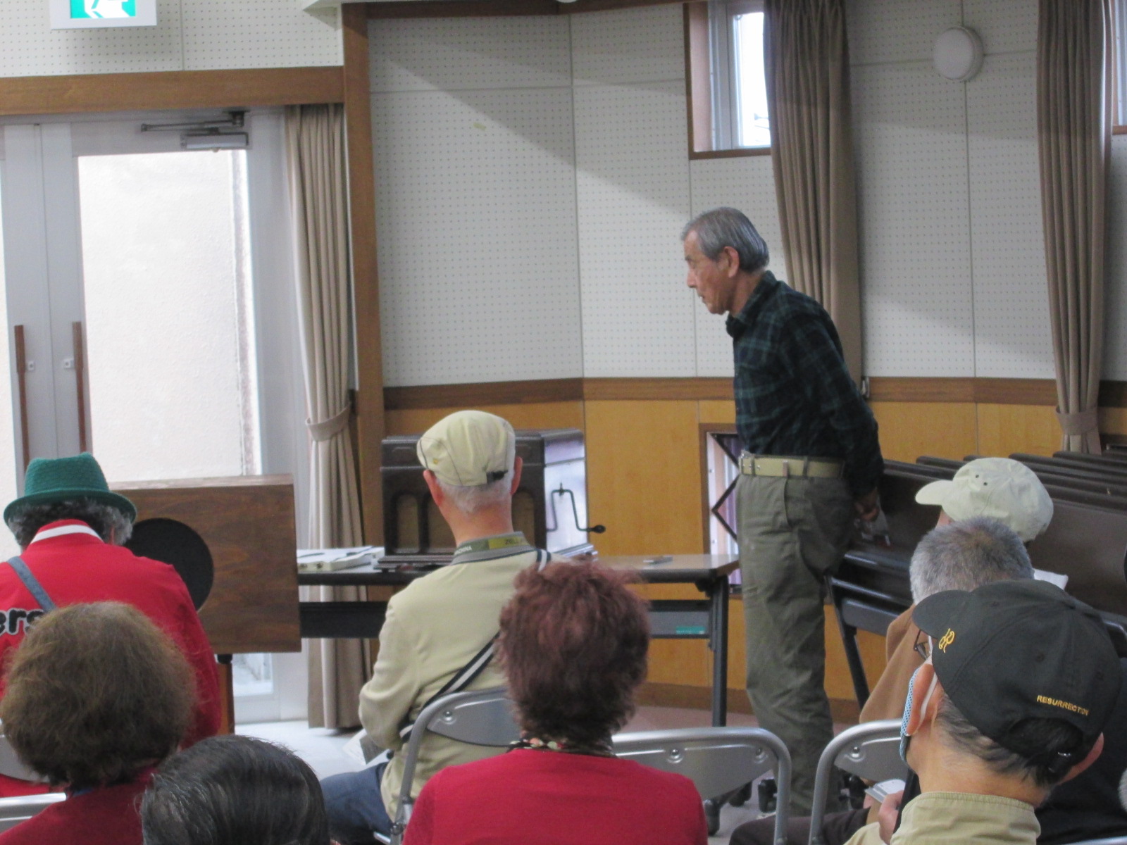 木目の腰板と白い壁の部屋で、後方の参加者に背中を向けている男性がテーブルの前に立っており、手前には数人の参加者が椅子に座っている様子の写真