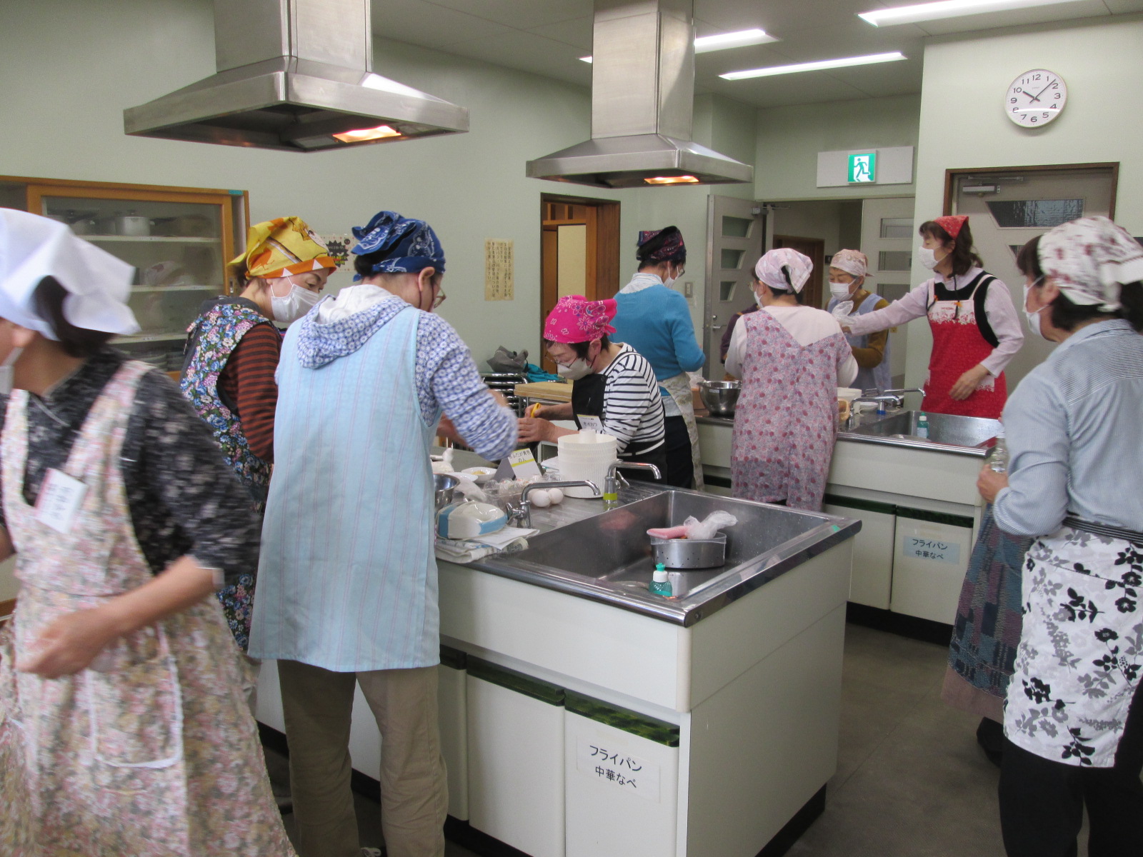 多くのエプロンとバンダナまたは三角巾を着用した女性たちが、ステンレス製の調理台と大きな換気フードがある清潔な調理室で、料理や製菓を楽しんでいる様子の写真