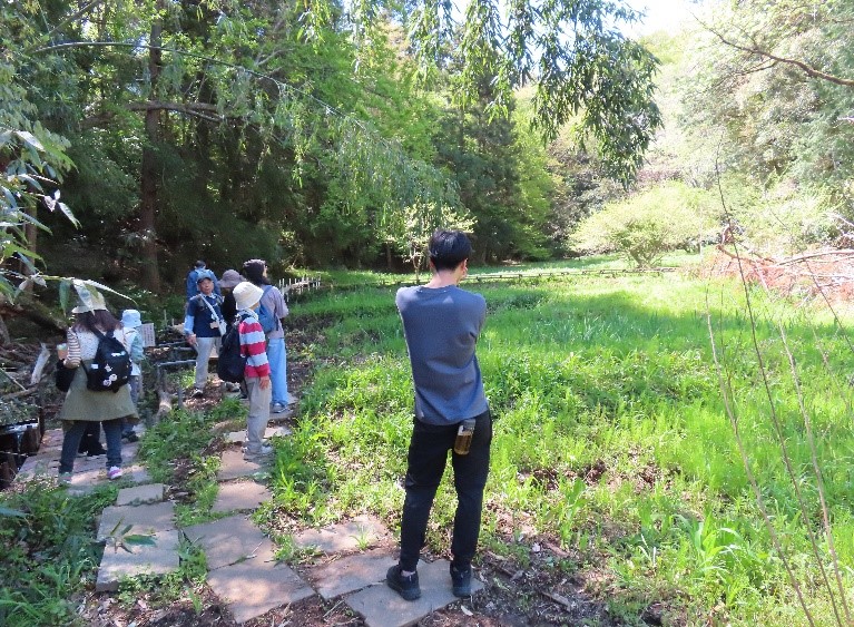 豊かな緑に囲まれた雑木林の中で、石畳の道を歩く人々と、カメラのようなものを構えて草地の広がる空間を観察している黒い服の男性を捉えた自然観察会の様子の写真