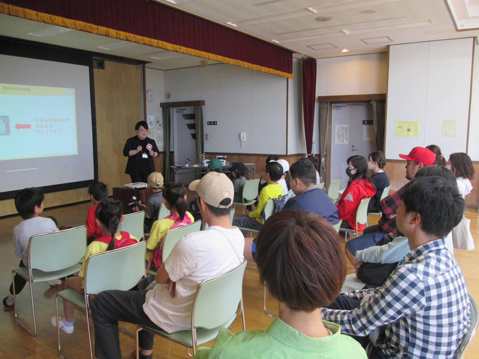 教室の前方で講師が話している中、子供たちと大人たちが椅子に座って静かに講義を聞いている様子を写した写真