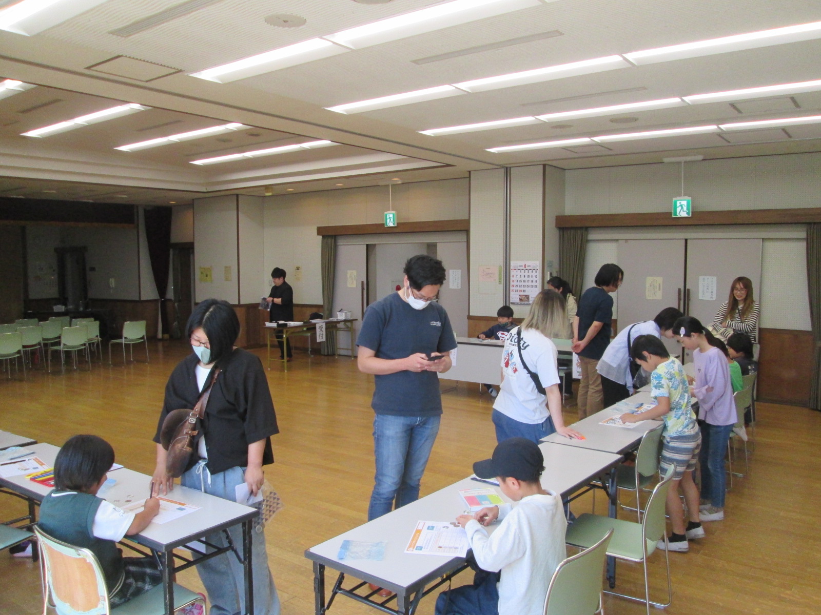 広い部屋の中で、子供たちが机に向かって作業をしており、周囲で大人たちが見守っている様子を写した写真