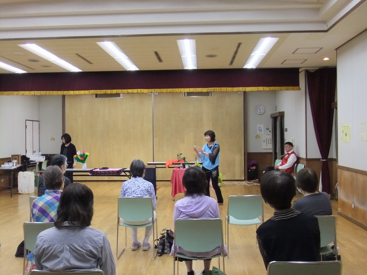 複数の参加者が着席する中、水色と黒の衣装を着た女性が前でマジックを披露している様子を捉えた写真