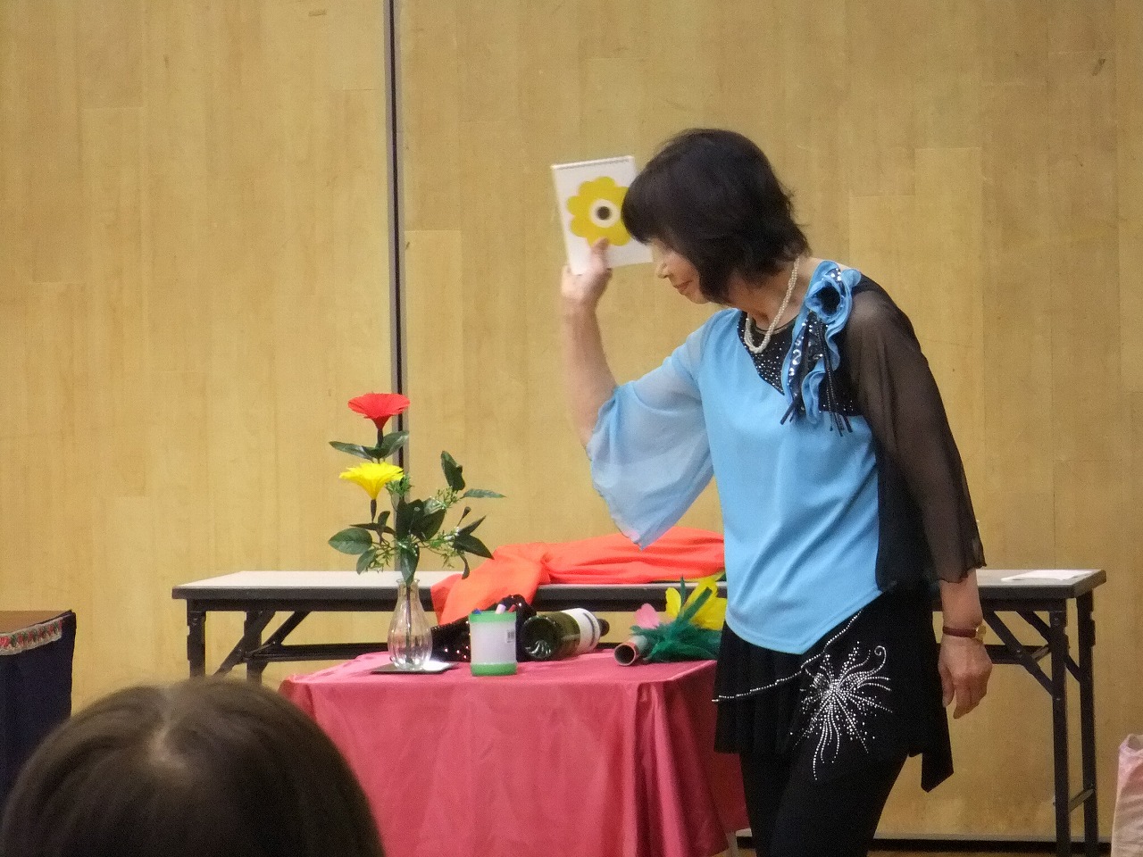 水色と黒の衣装を着た女性がテーブルの上の花や道具を使い、花柄のカードを見せながら観客の前で手品ショーをしている写真