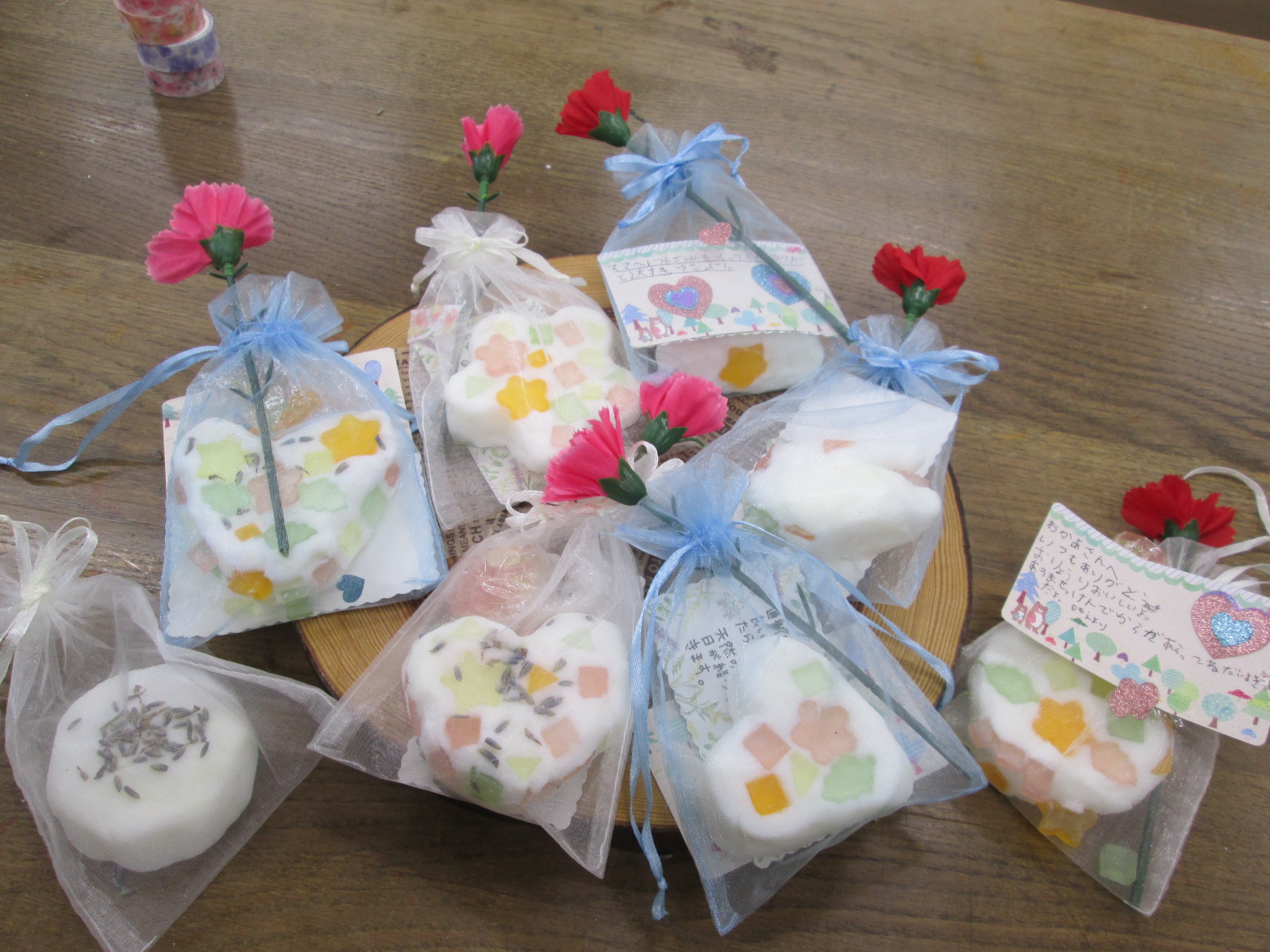 カラフルな小さなかけらを混ぜ込んだハート型や丸型の石鹸が、ピンクのカーネーションと青や白のリボンで飾られたオーガンジーの袋に入れられ、メッセージカードとともに並べられている手作りプレゼントの様子の写真