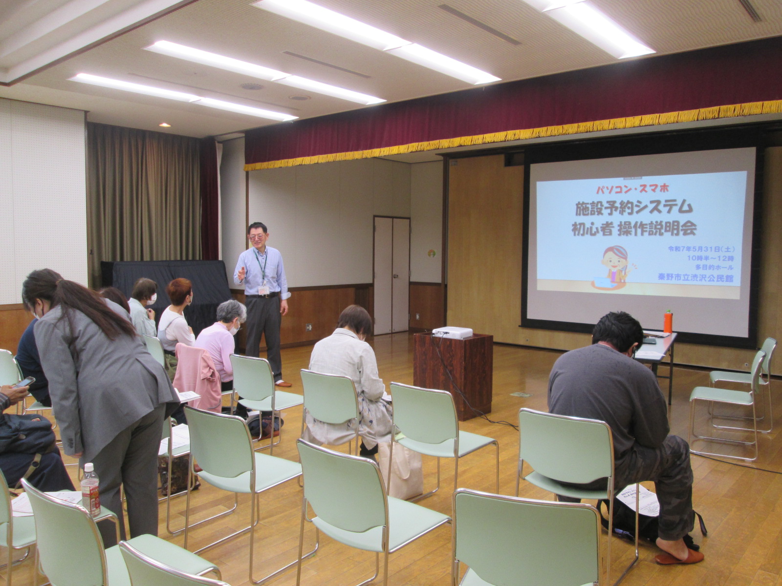 広い室内で数人が椅子に座り、プロジェクターに映し出されたパソコンやスマートフォンの操作説明を熱心に聞いている説明会の写真