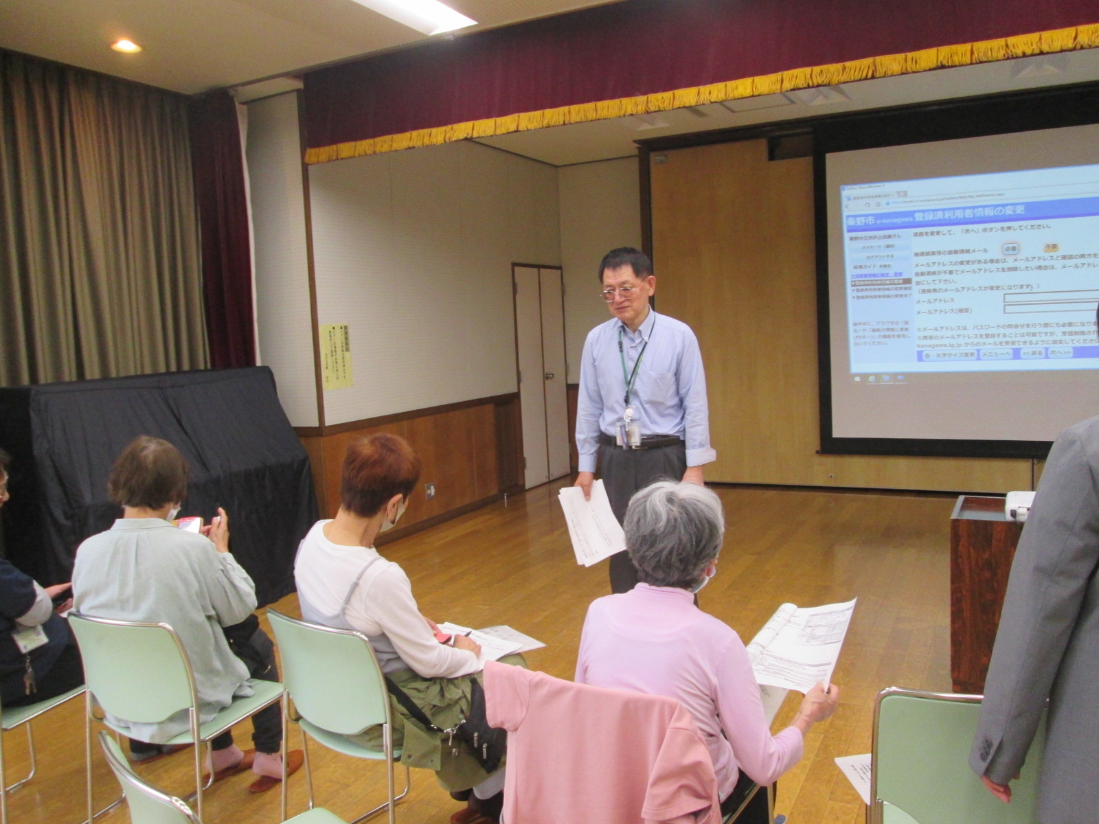 室内でプロジェクターに映し出された画面を背景に、ネームプレートをつけたシャツ姿の男性が数枚の資料を持ち、椅子に座っている参加者数人に説明を行っている写真