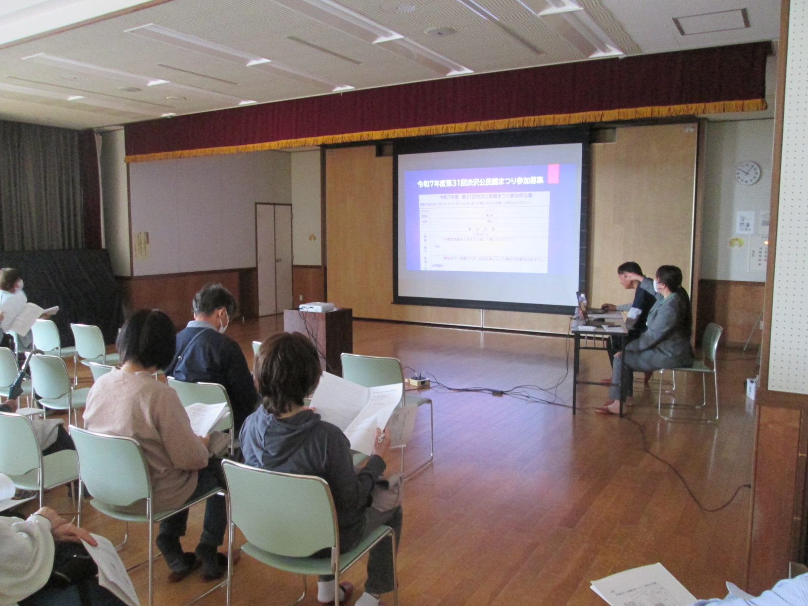 クリーンのプロジェクターに「令和元年度第31回公民館まつり事業報告」という文字を含む表が表示されており、数名の参加者が椅子に座り、スクリーン前のテーブルで関係者が説明を行っている様子の写真
