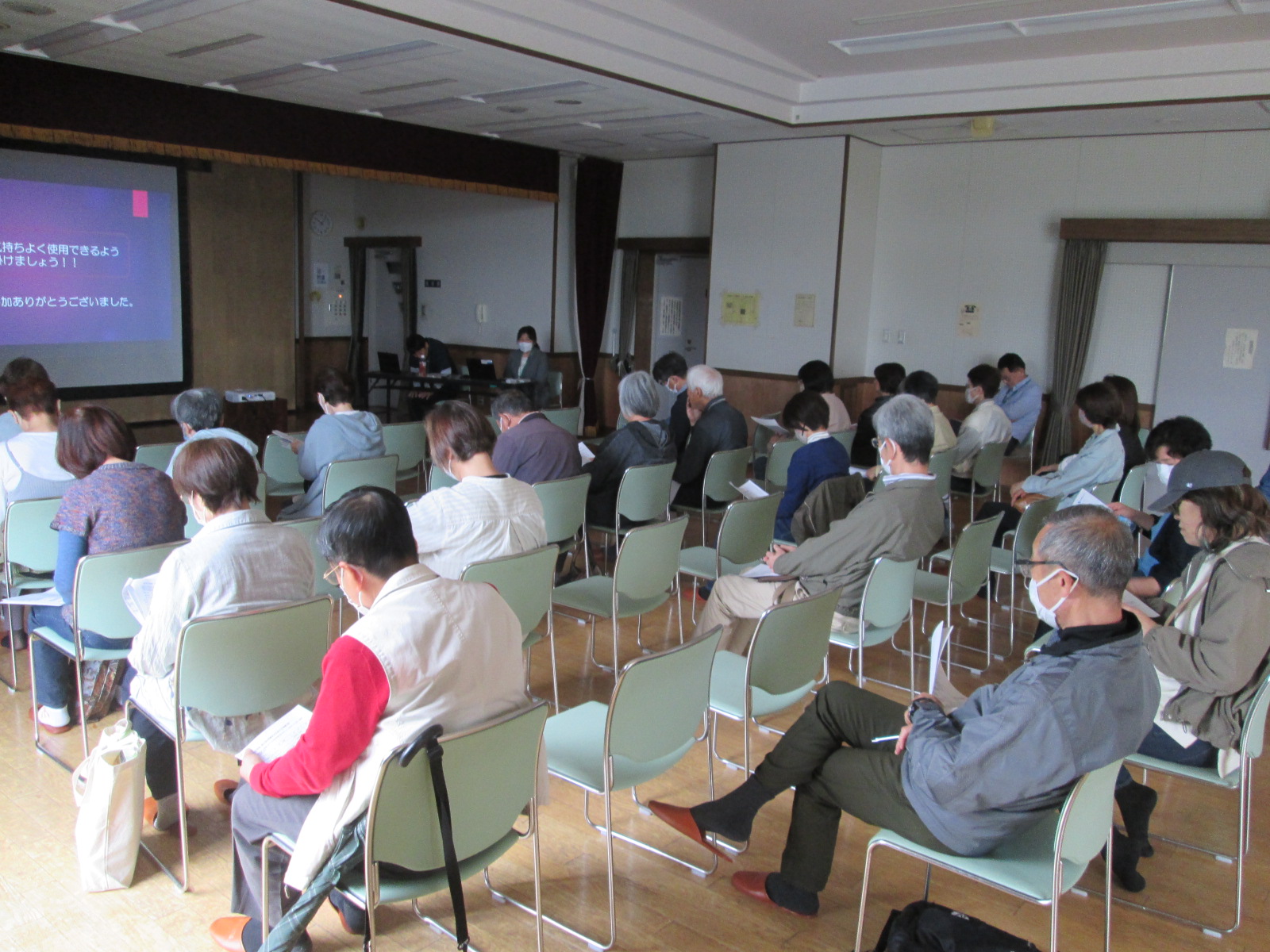 マスクを着用した多くの参加者が会場で緑色の椅子に座って前方のスクリーンに映し出された資料を静かに見つめている説明会の様子の写真