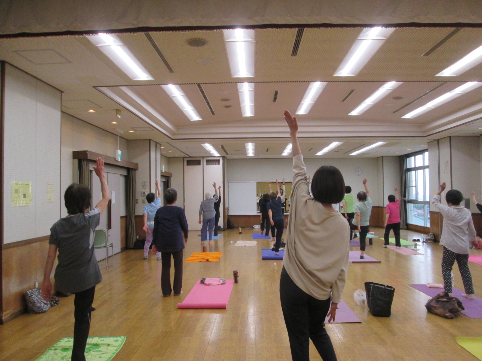 フローリングの広い室内で、十数人の人々が各自のヨガマットの上で、両腕を頭上に伸ばすポーズなどのストレッチやヨガを行っている様子の写真