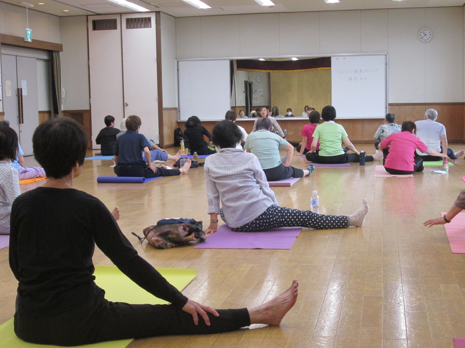 ヨガマットの上で体育座りや脚を伸ばすストレッチをするなど、高齢者や中高年の男女が室内で健康教室に参加している様子の写真