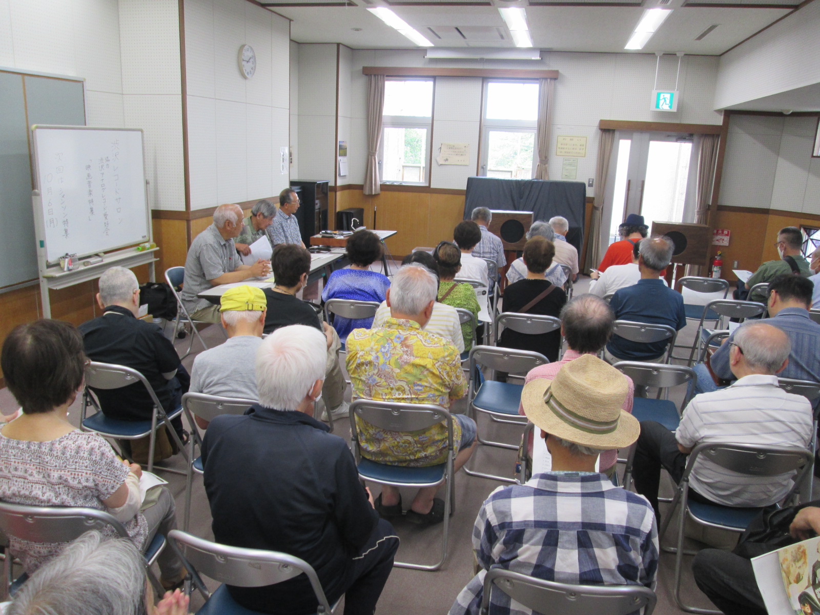 室内で多くの高齢者が奥のドアや窓を背にするように椅子に座り、左側のホワイトボードの前にいる数人の講師と思われる人物の話を聞いている写真
