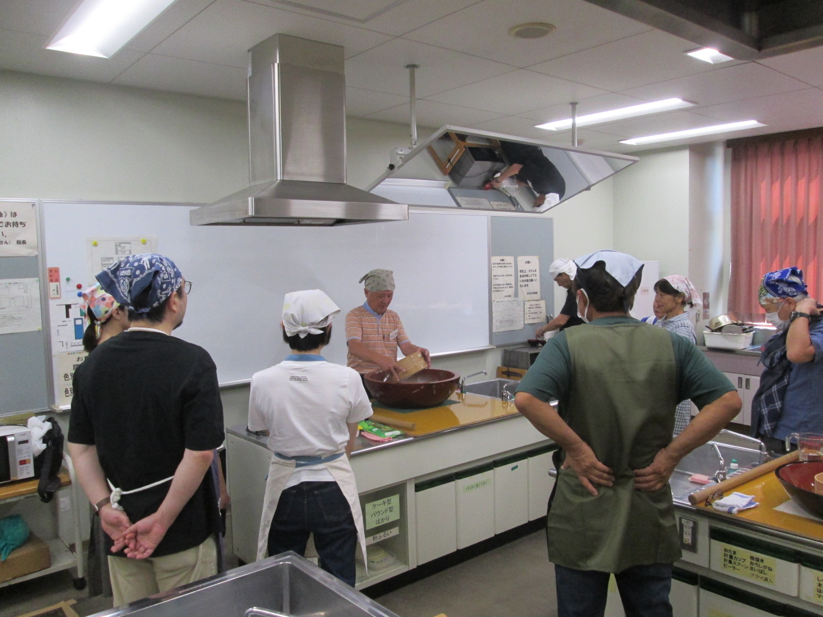 男女数人が頭にバンダナや三角巾をつけ、台所に立つ一人の男性から料理を教わっている様子の写真