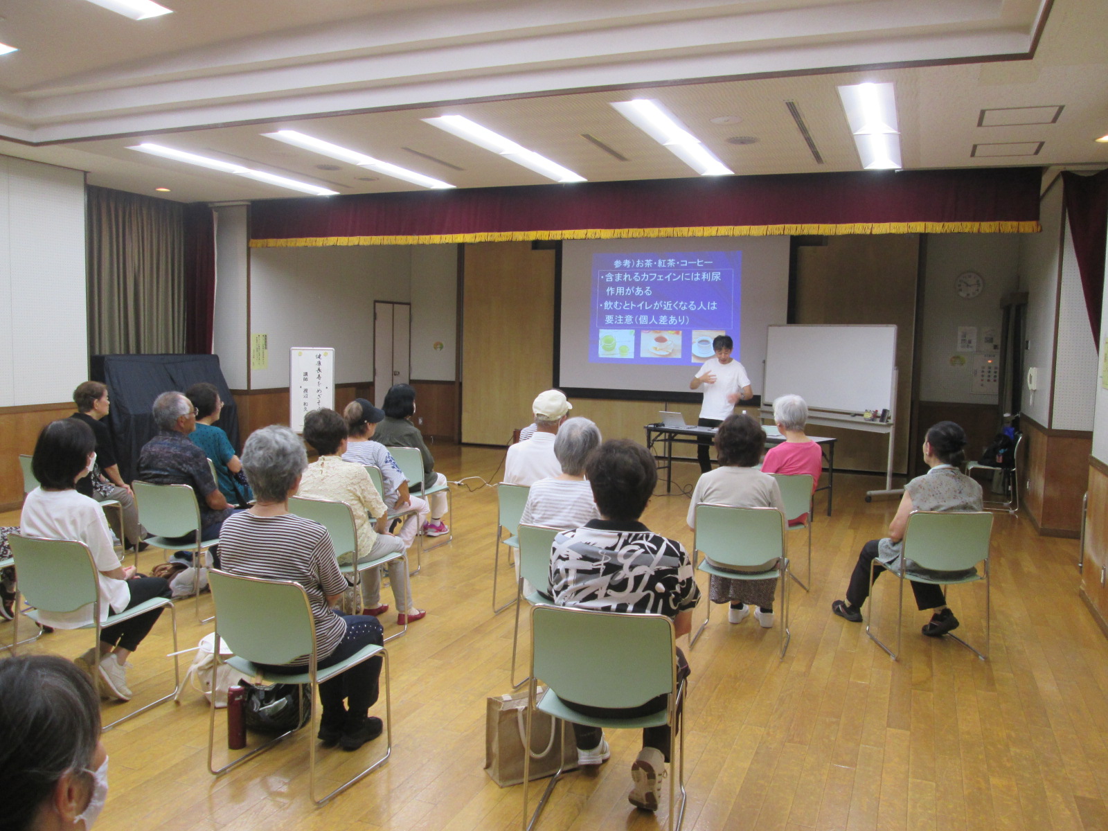室内で行われている健康講座で、渡邊先生がプロジェクターの画面を指しながら、椅子に座った高齢者たちに説明を行っている様子を捉えた写真