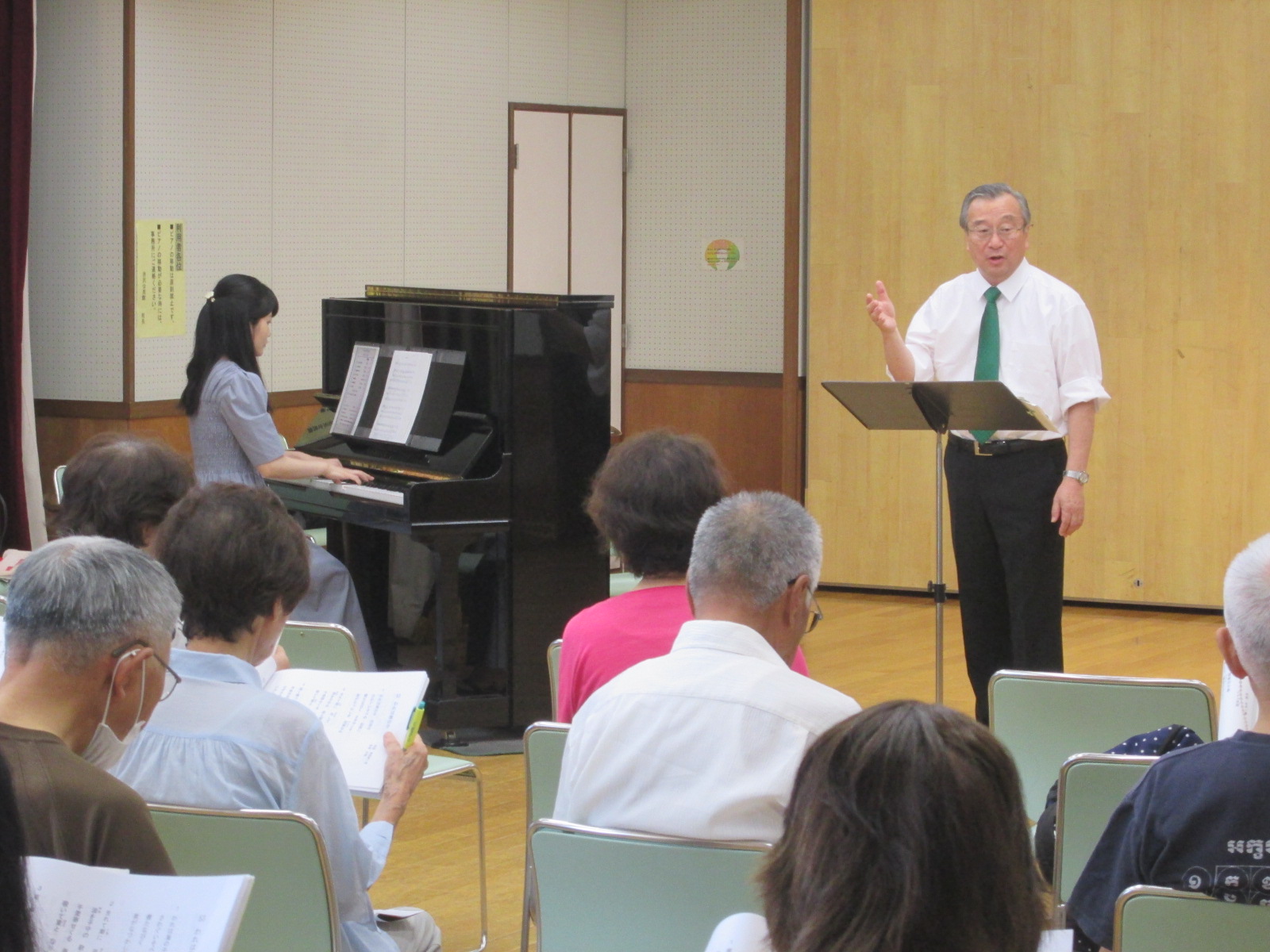 白いシャツに緑のネクタイを締めた男性が、木目調の壁を背景に指揮台の前で聴衆に向かって話しており、左奥では女性が黒いアップライトピアノを弾き、数人の高齢者が座って歌詞を見ている合唱の様子の写真