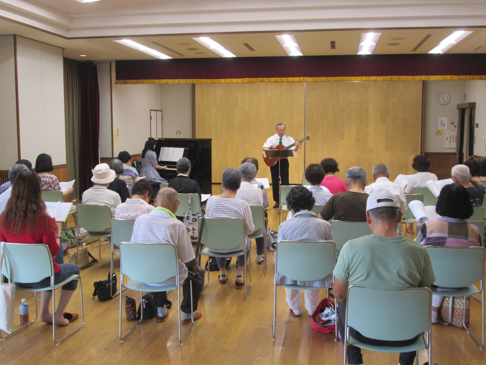 白いシャツに緑のネクタイの男性がギターを弾きながら歌を披露し、黒いピアノを弾く女性と多くの聴衆が緑色の椅子に座って鑑賞している様子の写真