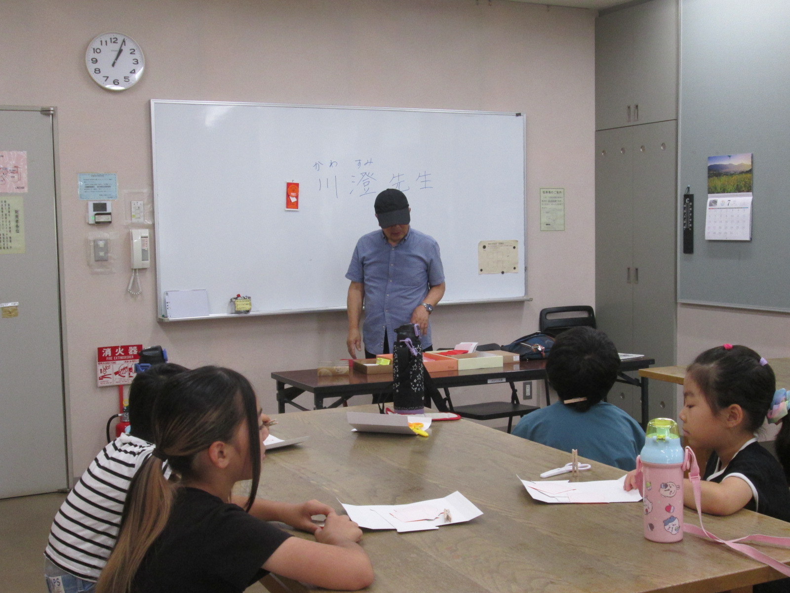 帽子をかぶった男性が、白いボードに「 川澄先生」と書かれた教室で、机に向かって座っている数人の子どもたちに対し何かを説明している様子の写真