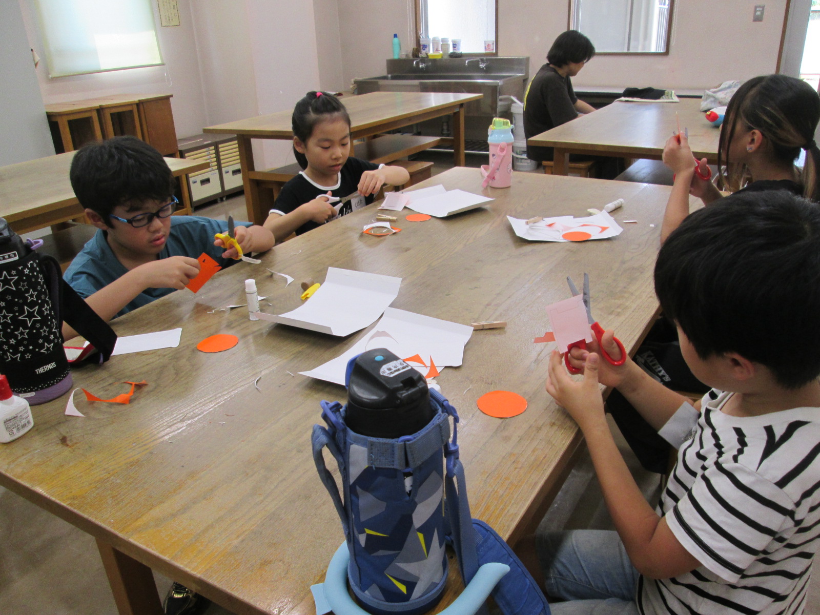 数人の子供たちが机を囲んで座り、紙をハサミで切ったり、糊で貼ったりして工作している様子の写真